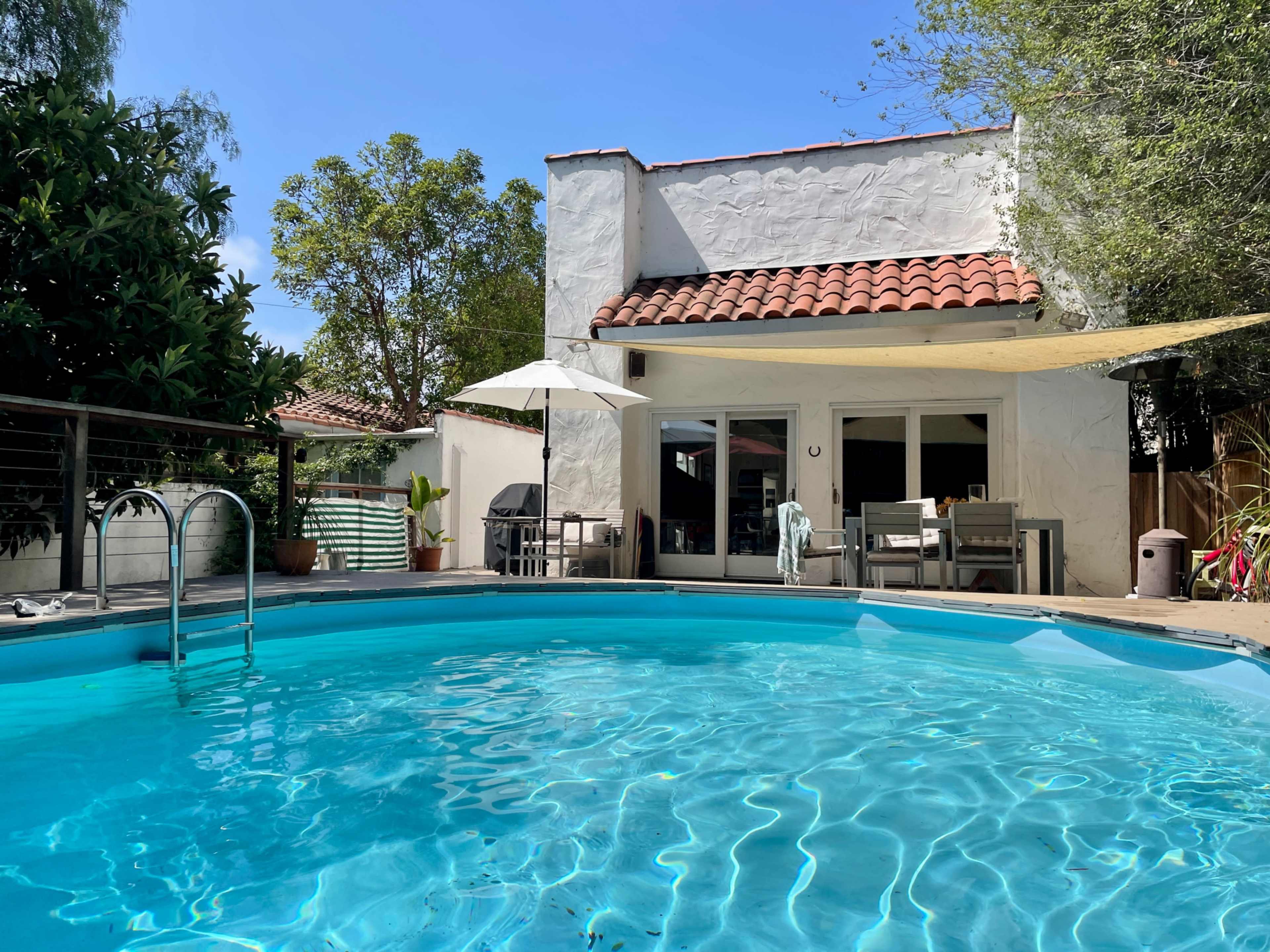 A circular swimming pool is surrounded by a patio and a small house with a tiled roof and large windows.
