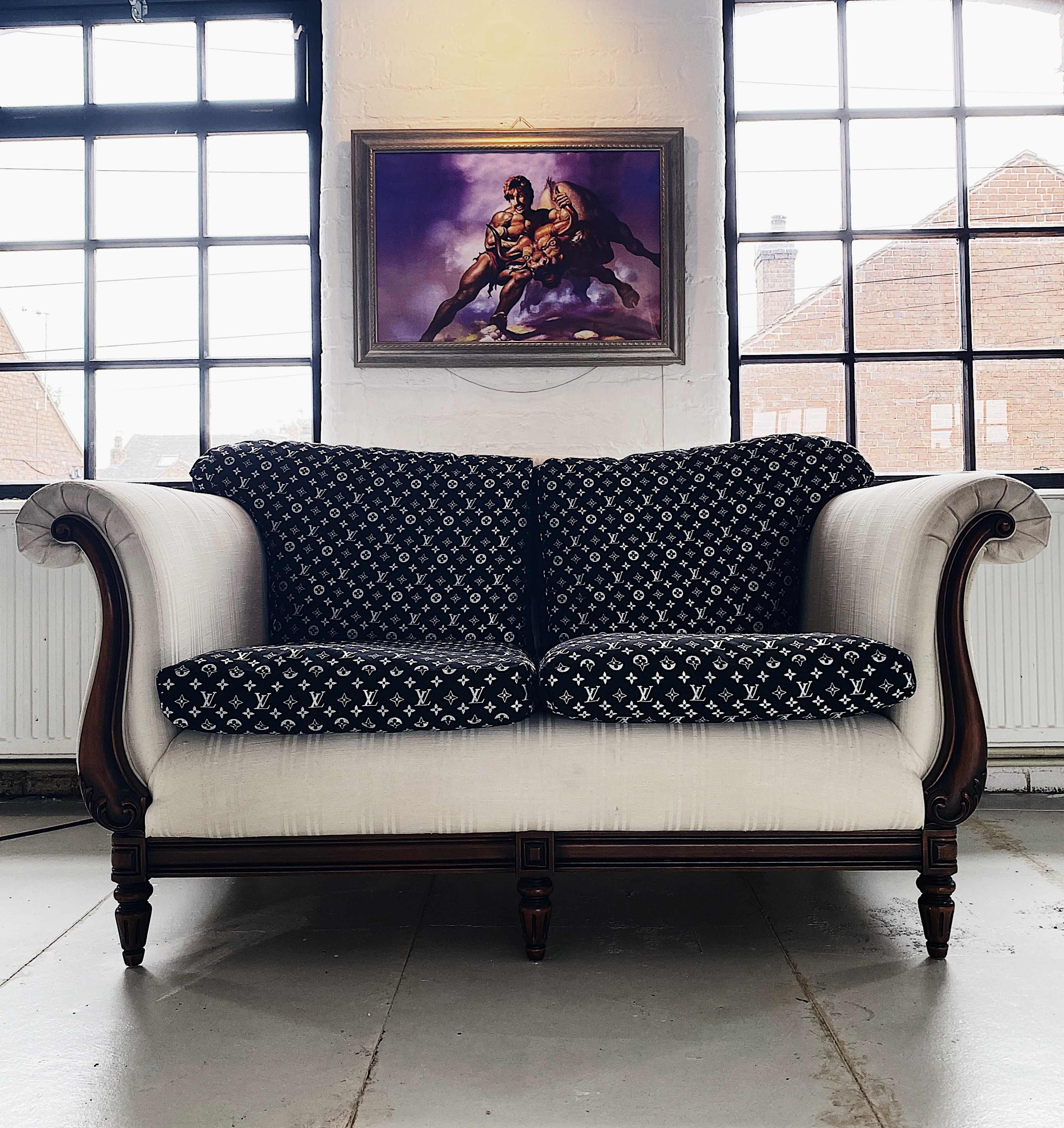A vintage two-seater sofa with a black patterned upholstery and white fabric, set against a wall with large windows and a framed painting above it.