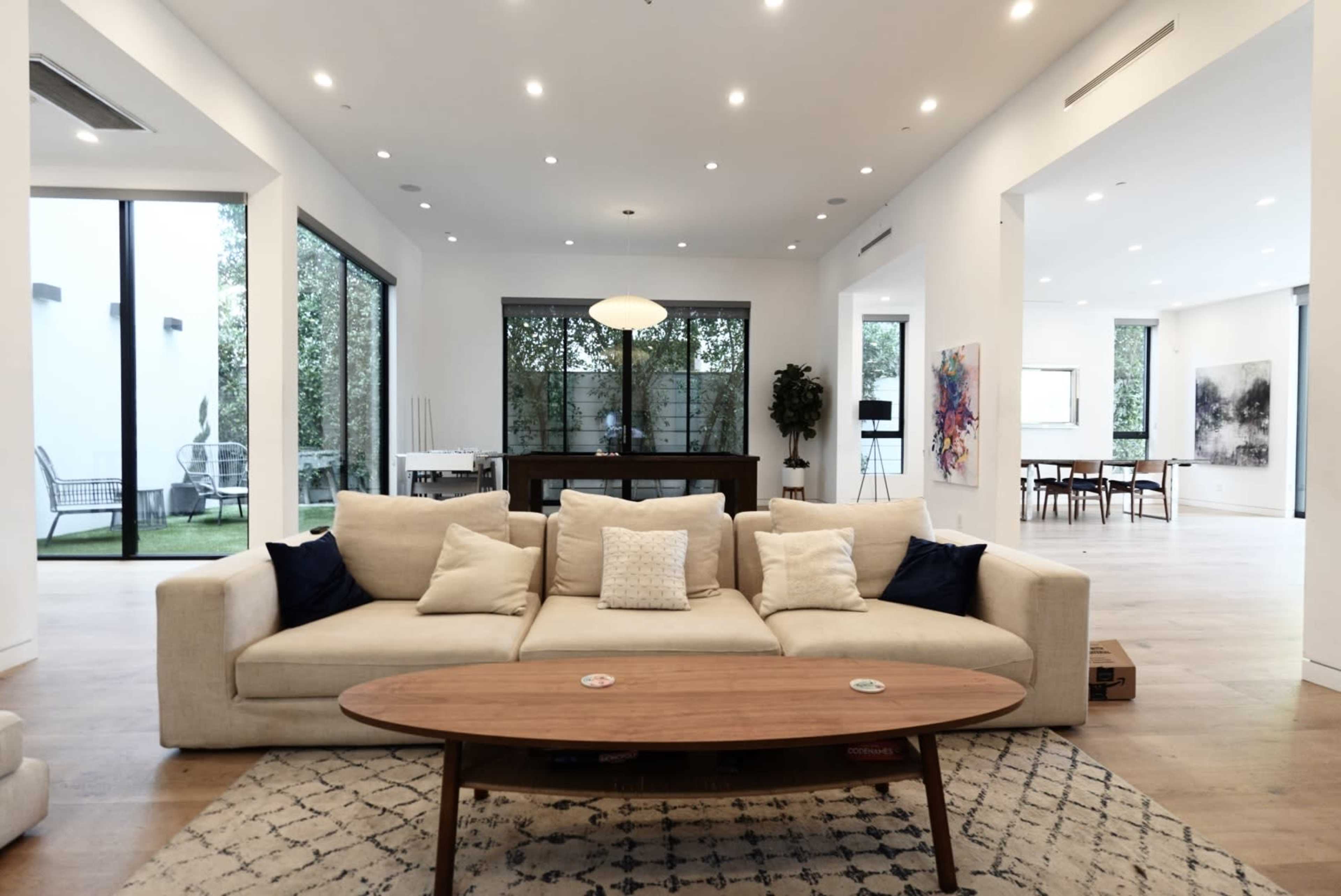 A modern living room features a beige sofa with decorative pillows and a wooden coffee table, overlooking a spacious dining area through large glass windows.