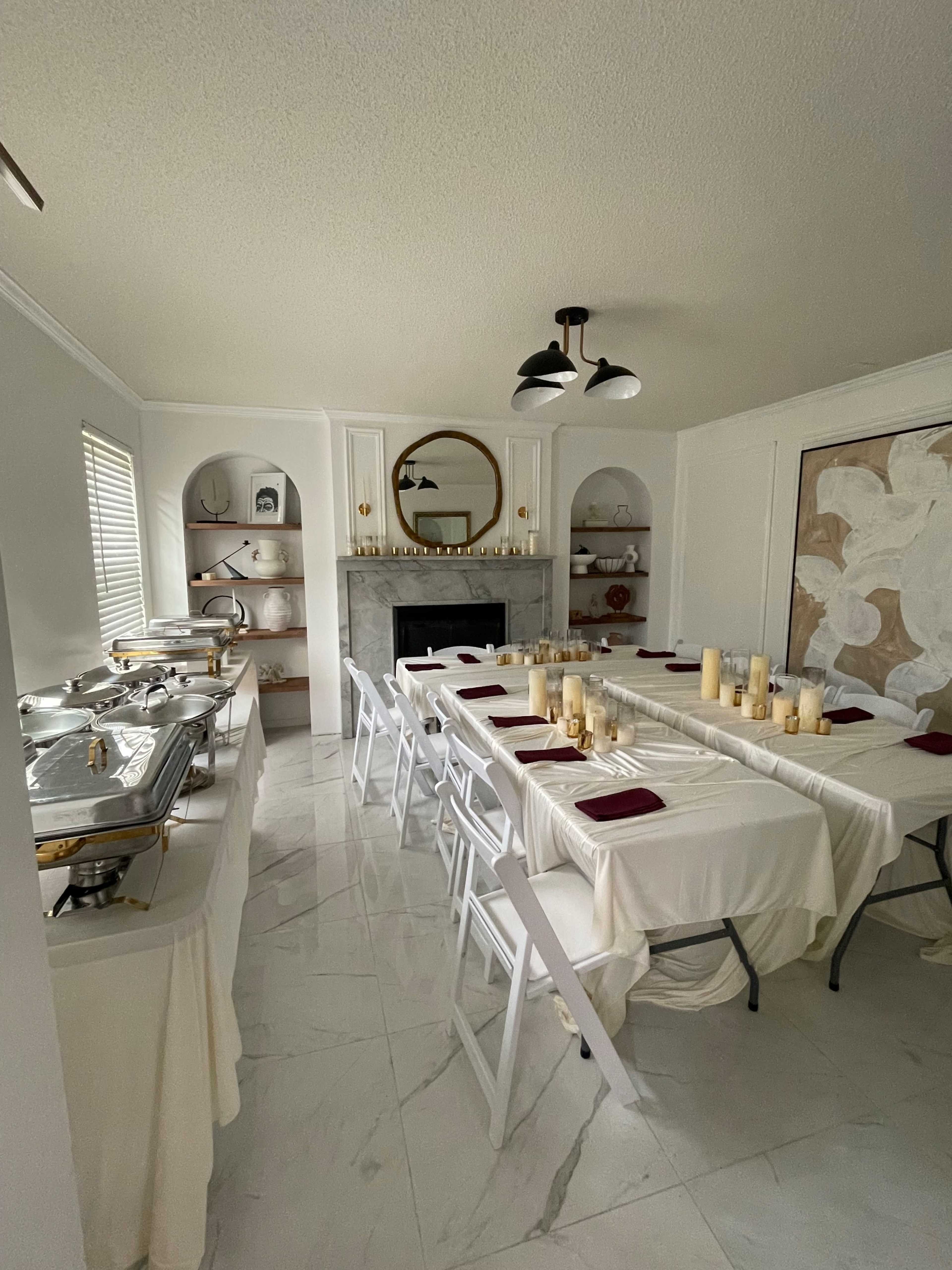 A dining room is set with a long table adorned with candles and cloths, facing a marble fireplace and shelves displaying decorative items.