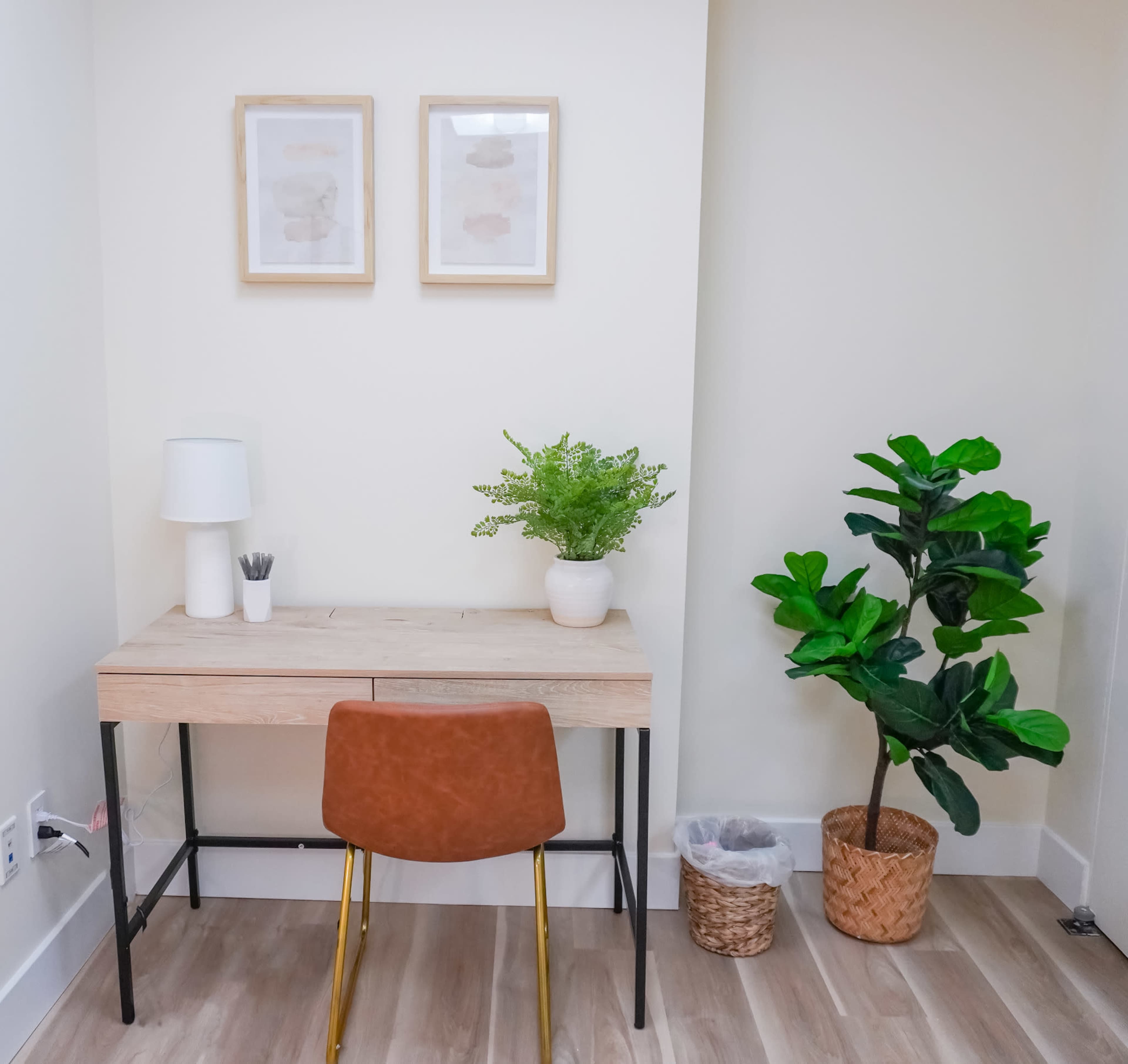 The image depicts a small home office space featuring a wooden desk with a brown chair, a lamp, potted plants, and framed artwork on the wall.