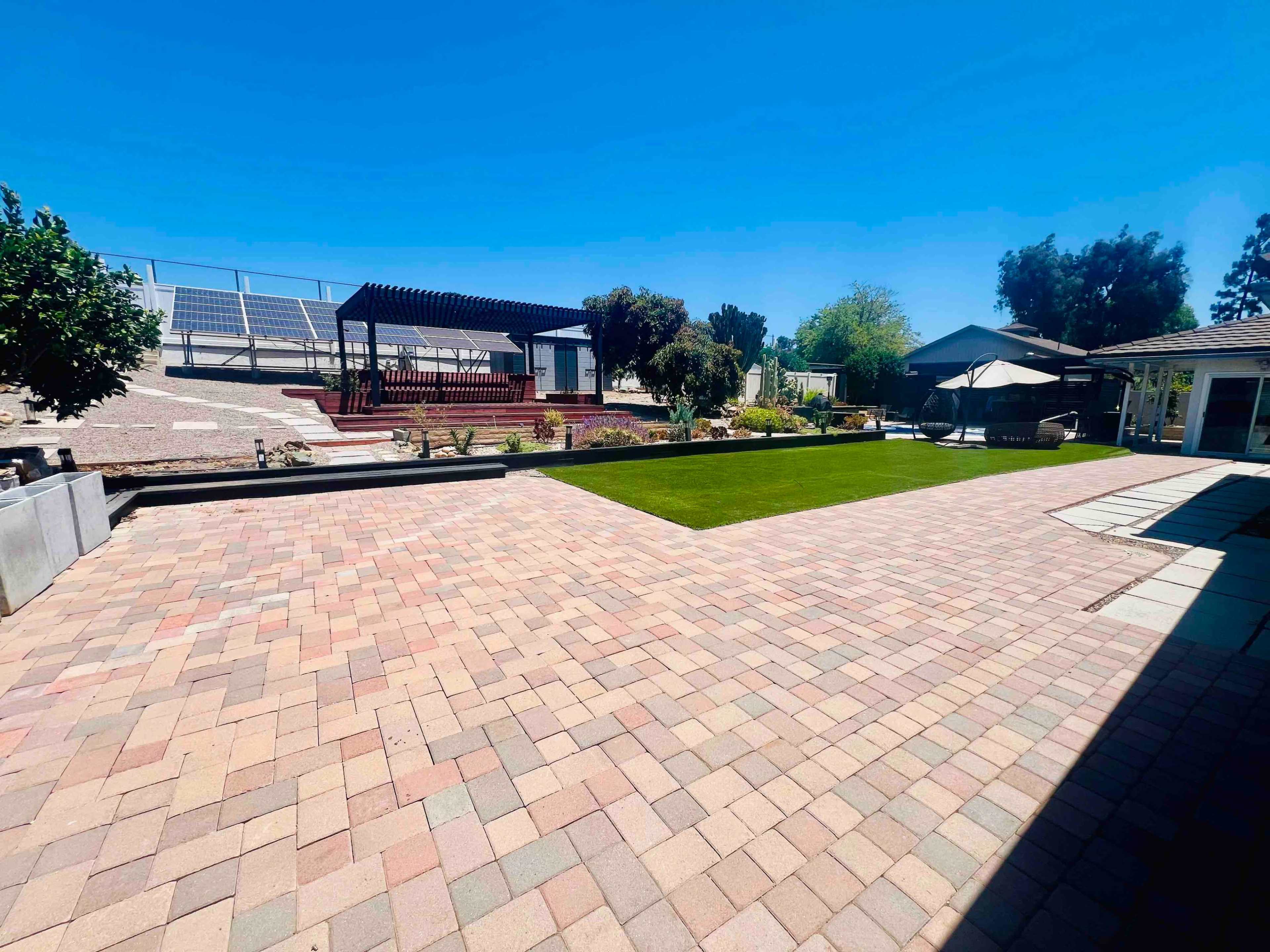The image shows a spacious backyard with a patio made of stone pavers, a pergola, landscaping features, and solar panels in the background.