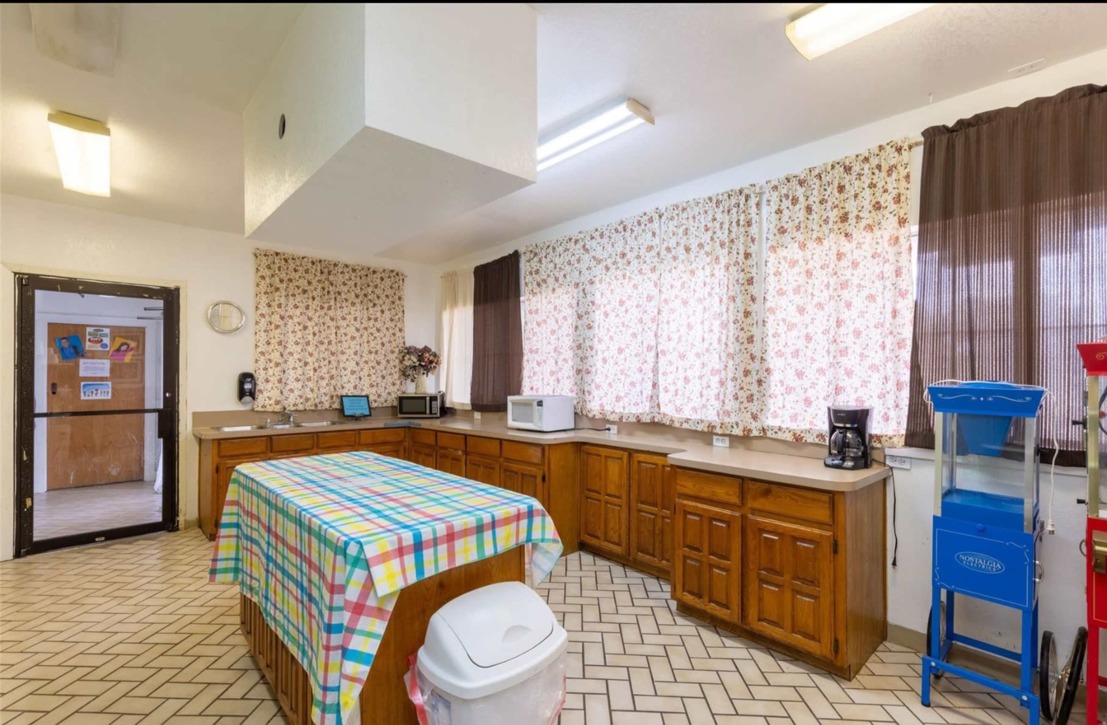 The image depicts a spacious kitchen with wooden cabinets, a checkered tablecloth covering an island, and large windows draped with floral curtains.