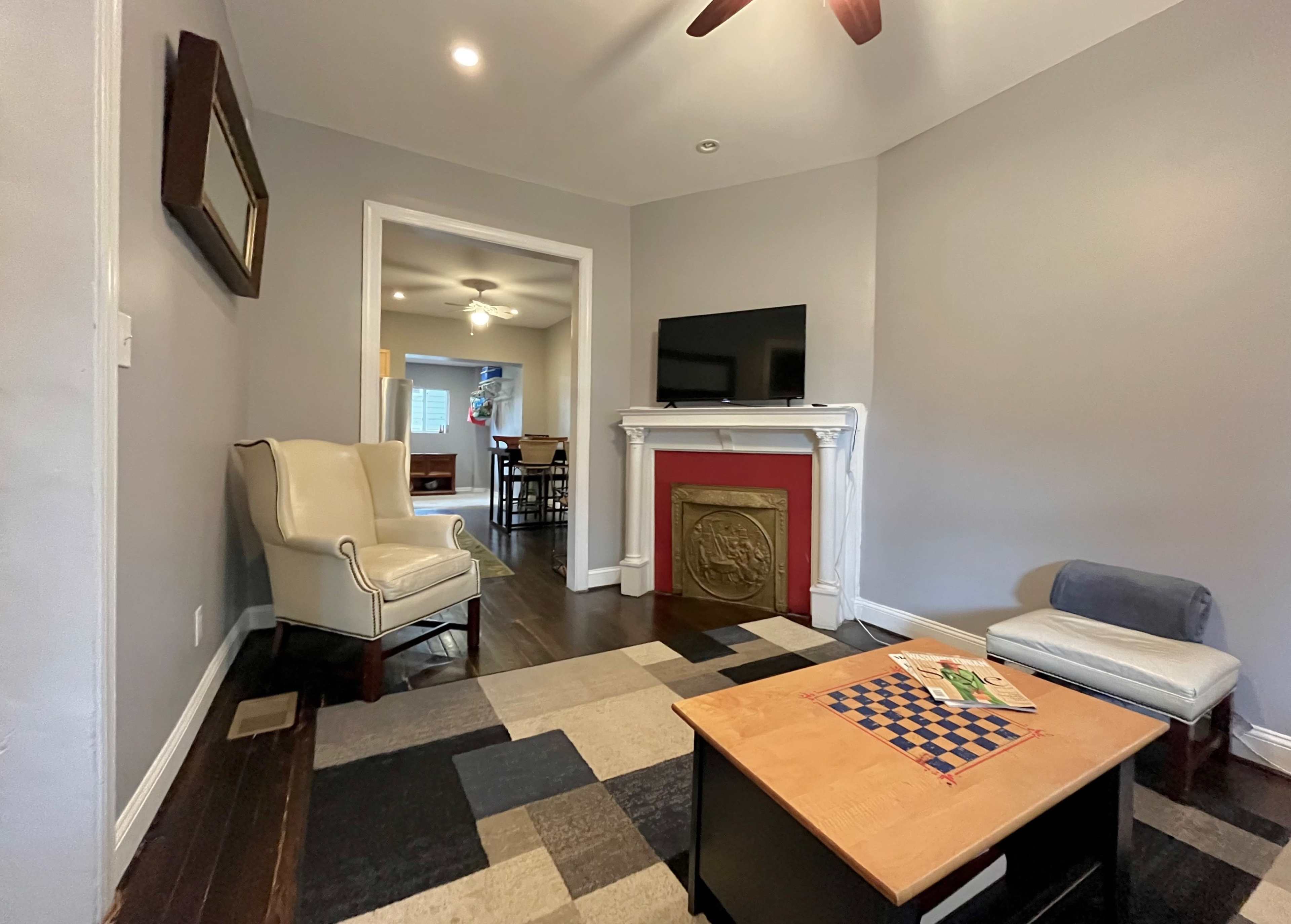 A living room with a white armchair, a flat-screen TV mounted on the wall, and a coffee table on a patterned rug.