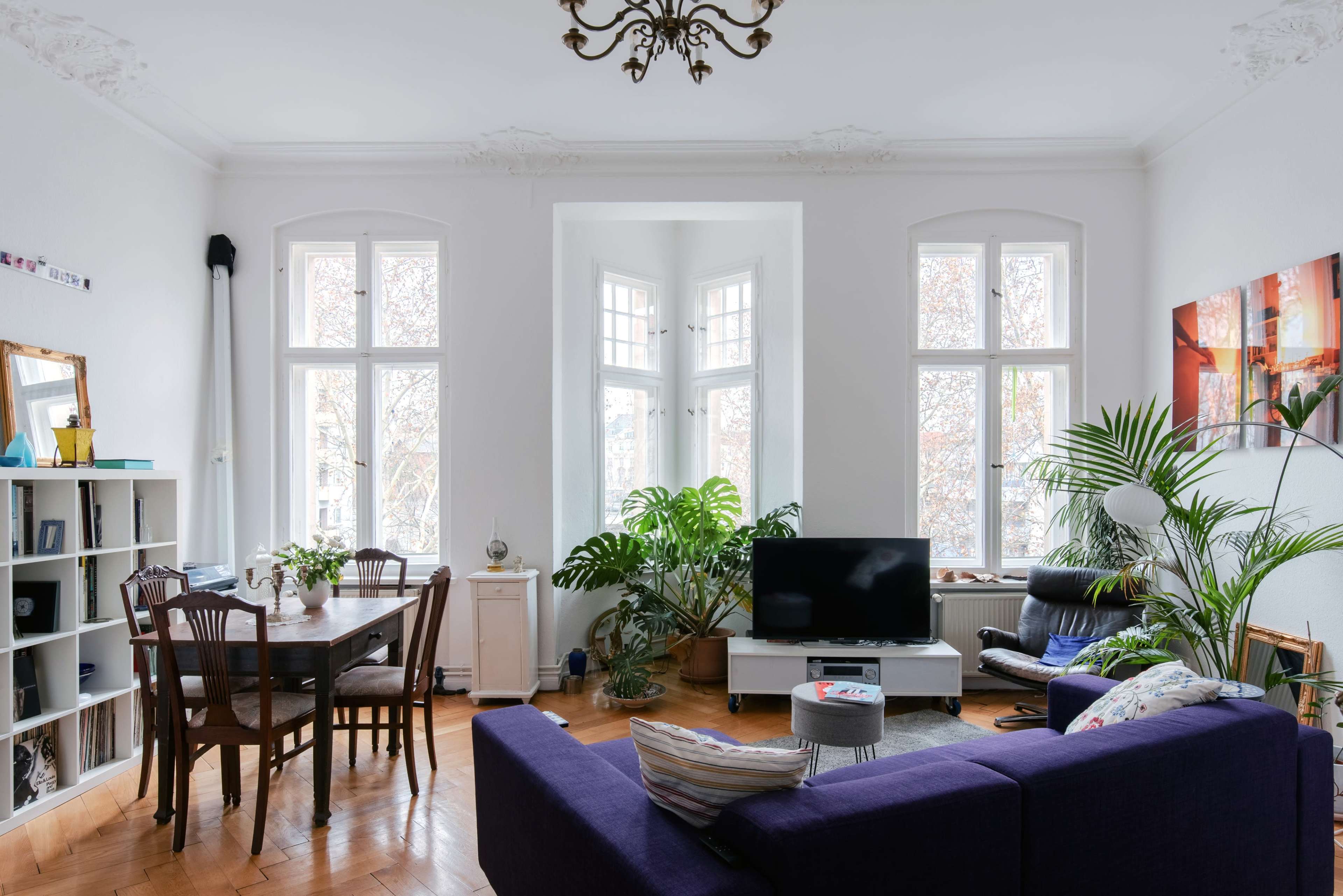 The image depicts a bright living room featuring a large window nook, a dining table, a television, and various plants.