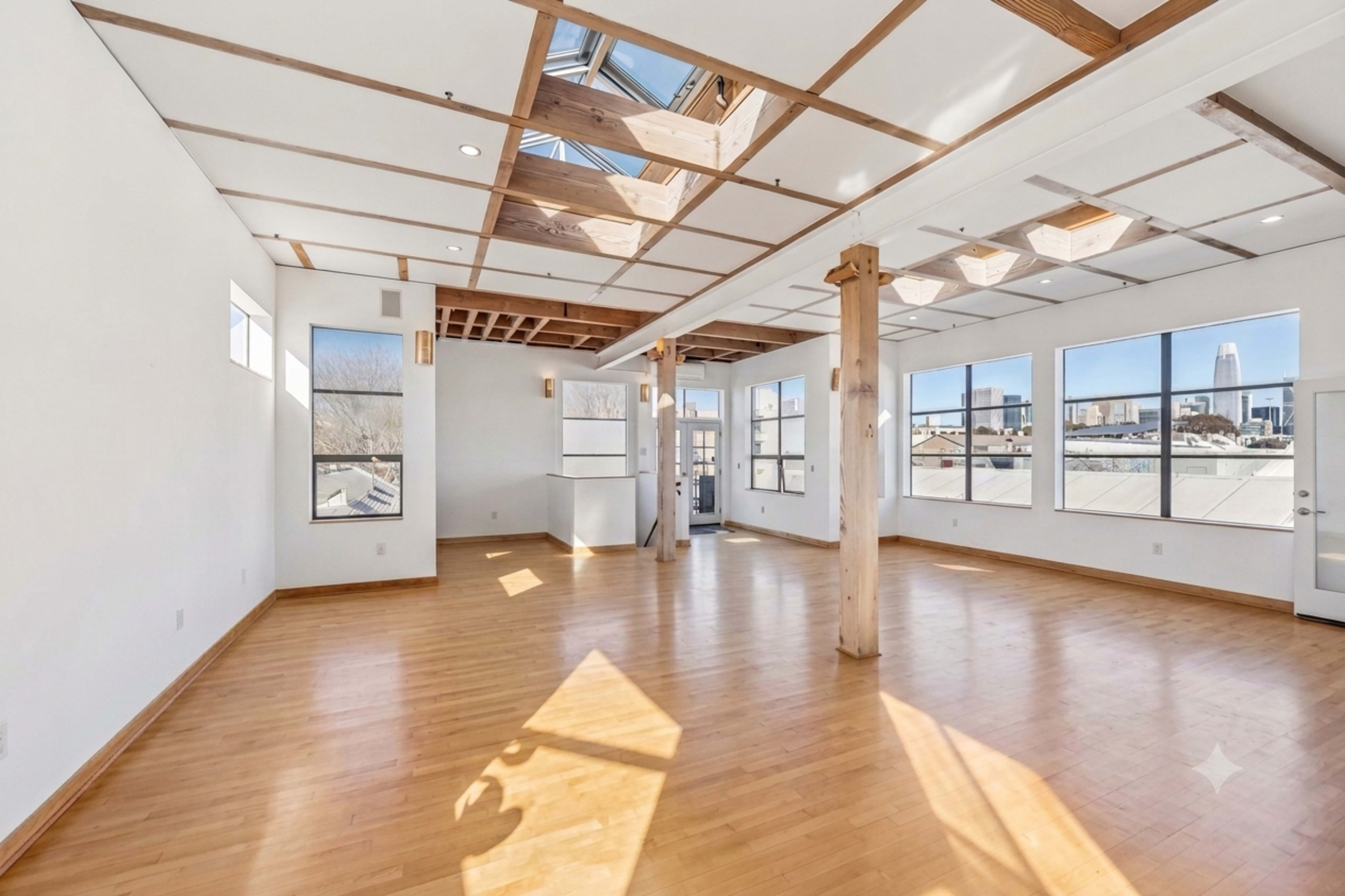 The image shows a spacious, well-lit room with wooden flooring, large windows, and a ceiling featuring exposed beams and skylights.