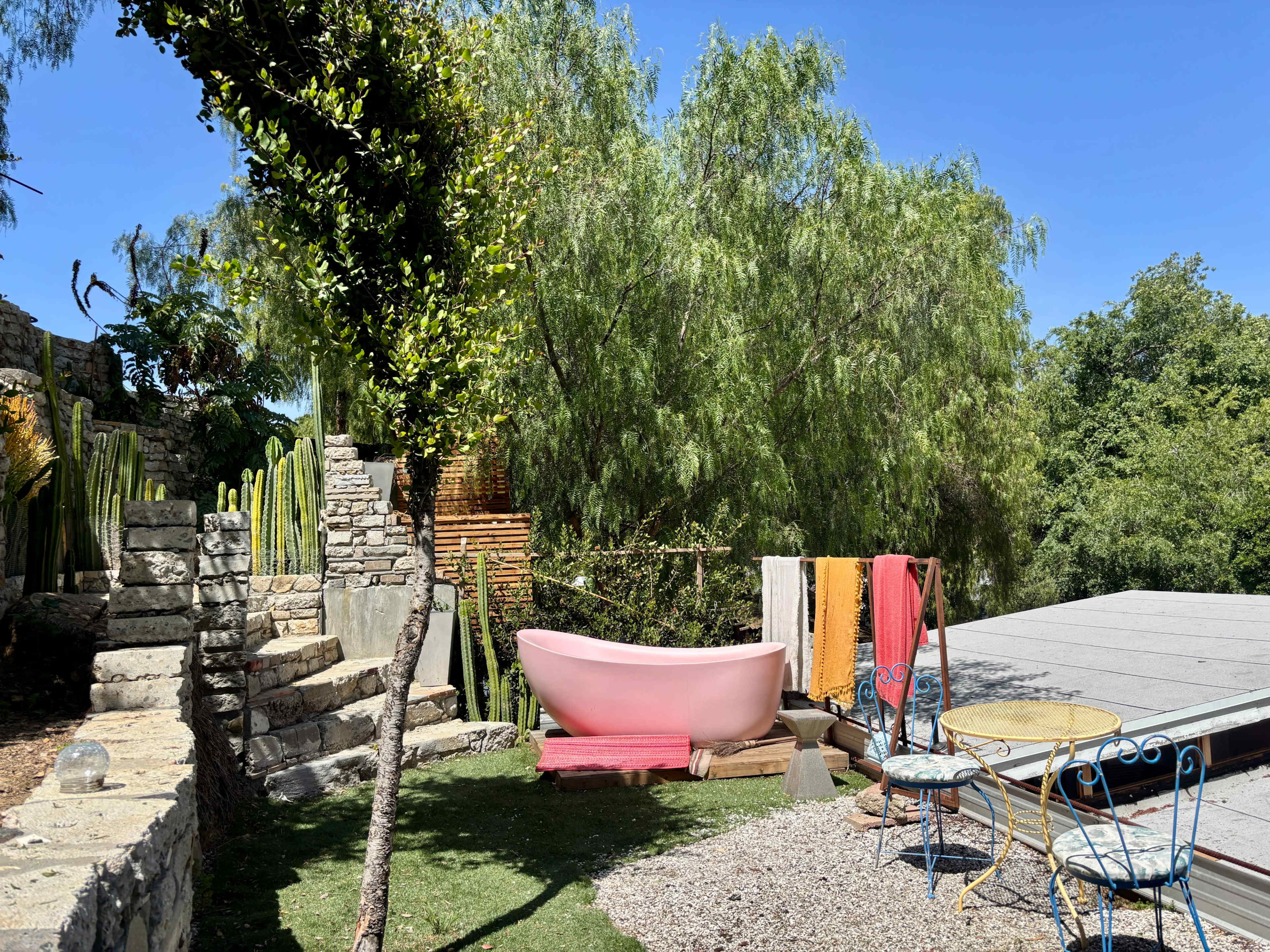 The scene features a pink bathtub surrounded by greenery, a stone pathway, and colorful textiles near a seating area with a small table and chairs.