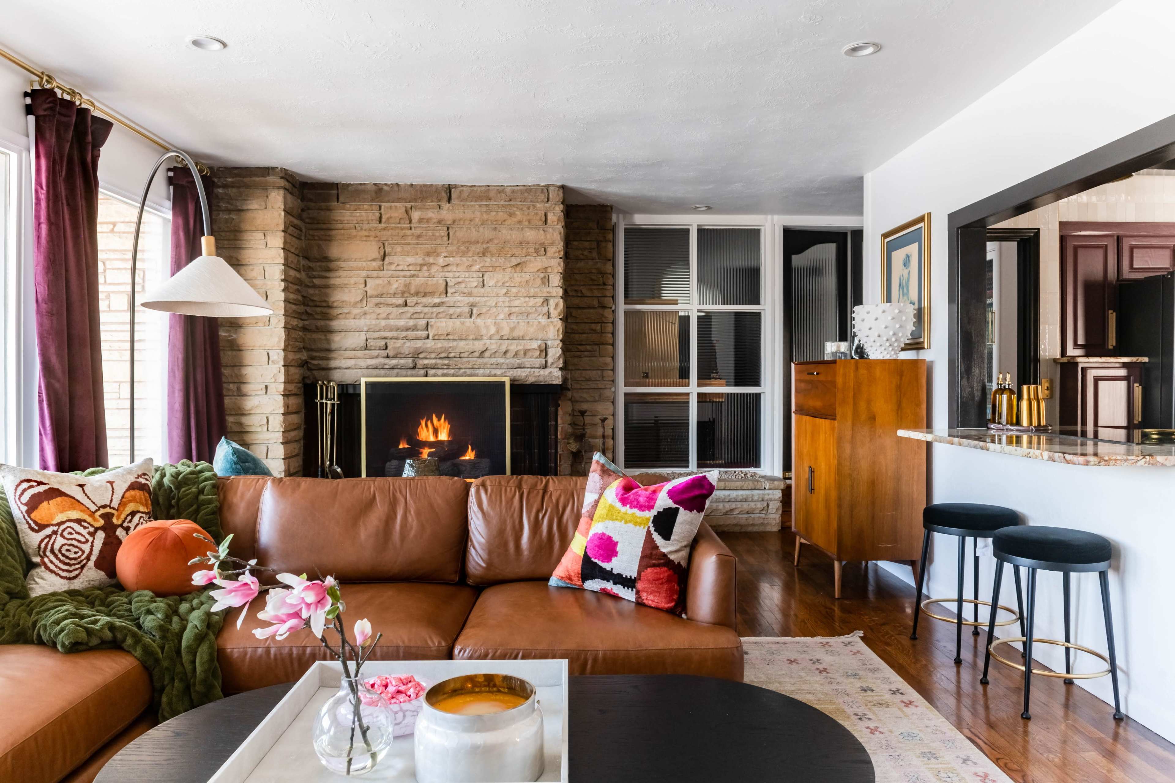 A cozy living room features a brown leather sectional sofa adorned with colorful pillows, a modern coffee table, and a stone fireplace, with a view of an open kitchen area.