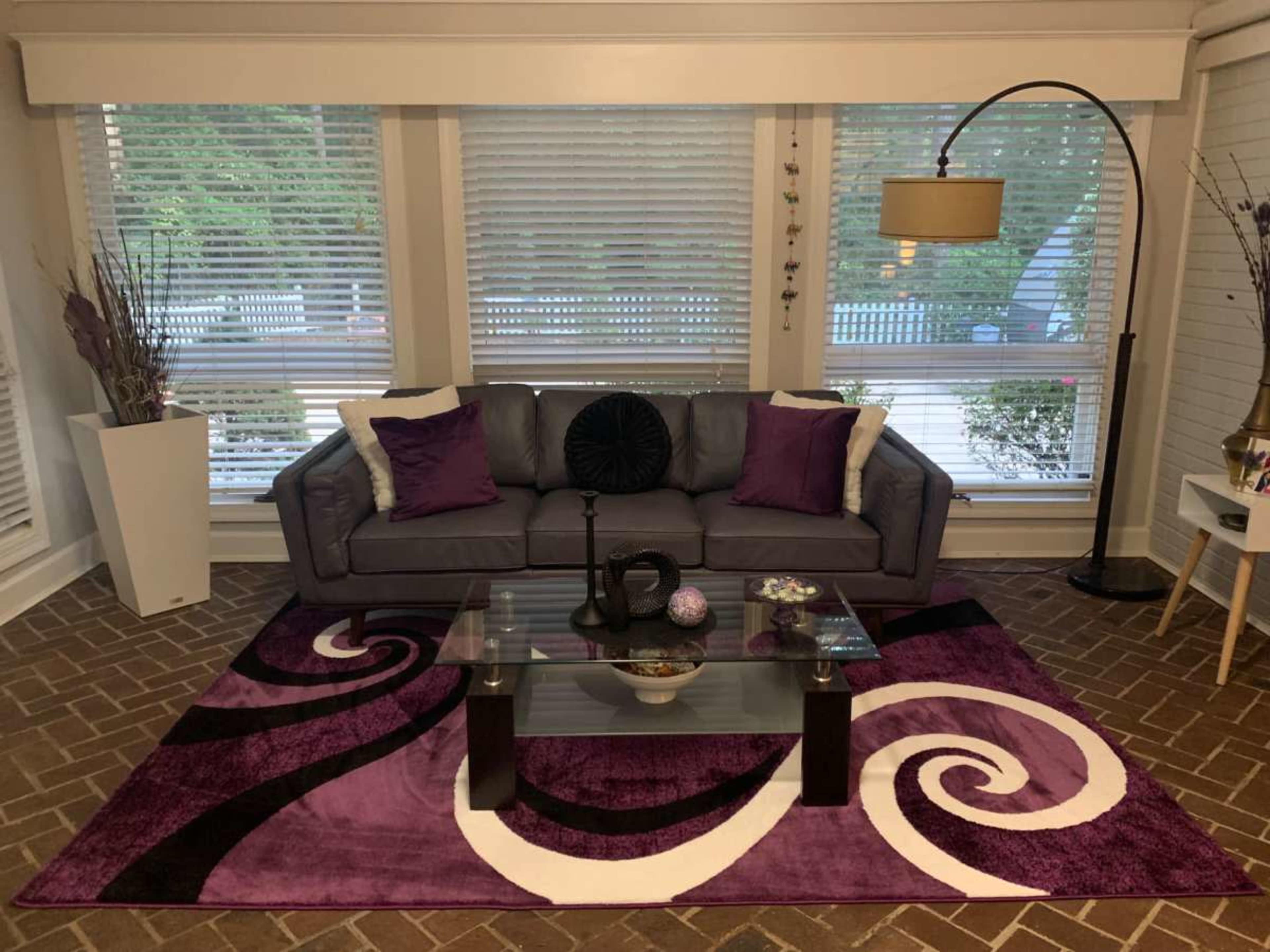 The living room features a gray sofa with purple cushions, a glass coffee table on a patterned purple rug, and large windows with blinds.