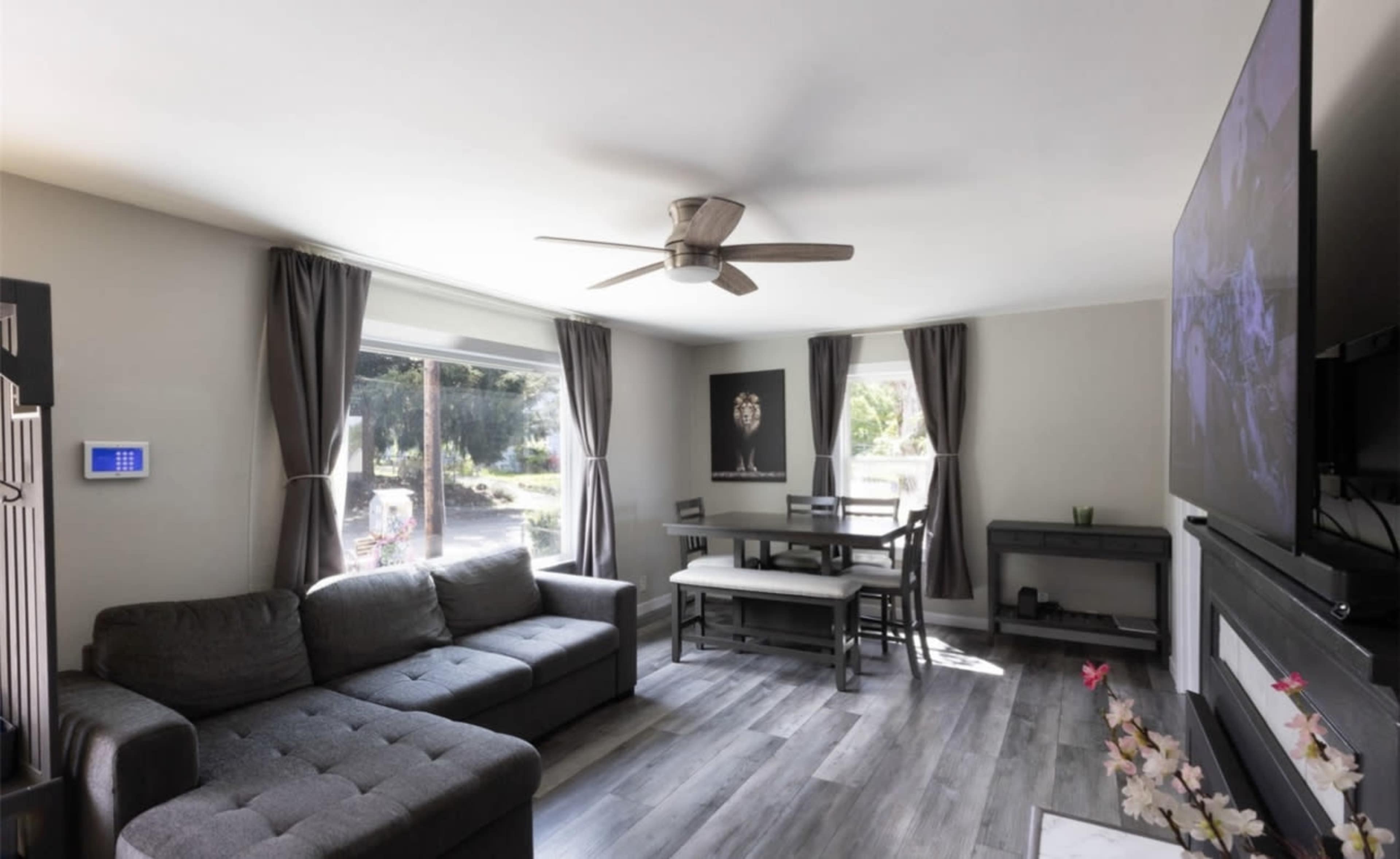 The image shows a modern living room with a gray sectional sofa, a dining table with chairs, and large windows bringing in natural light.