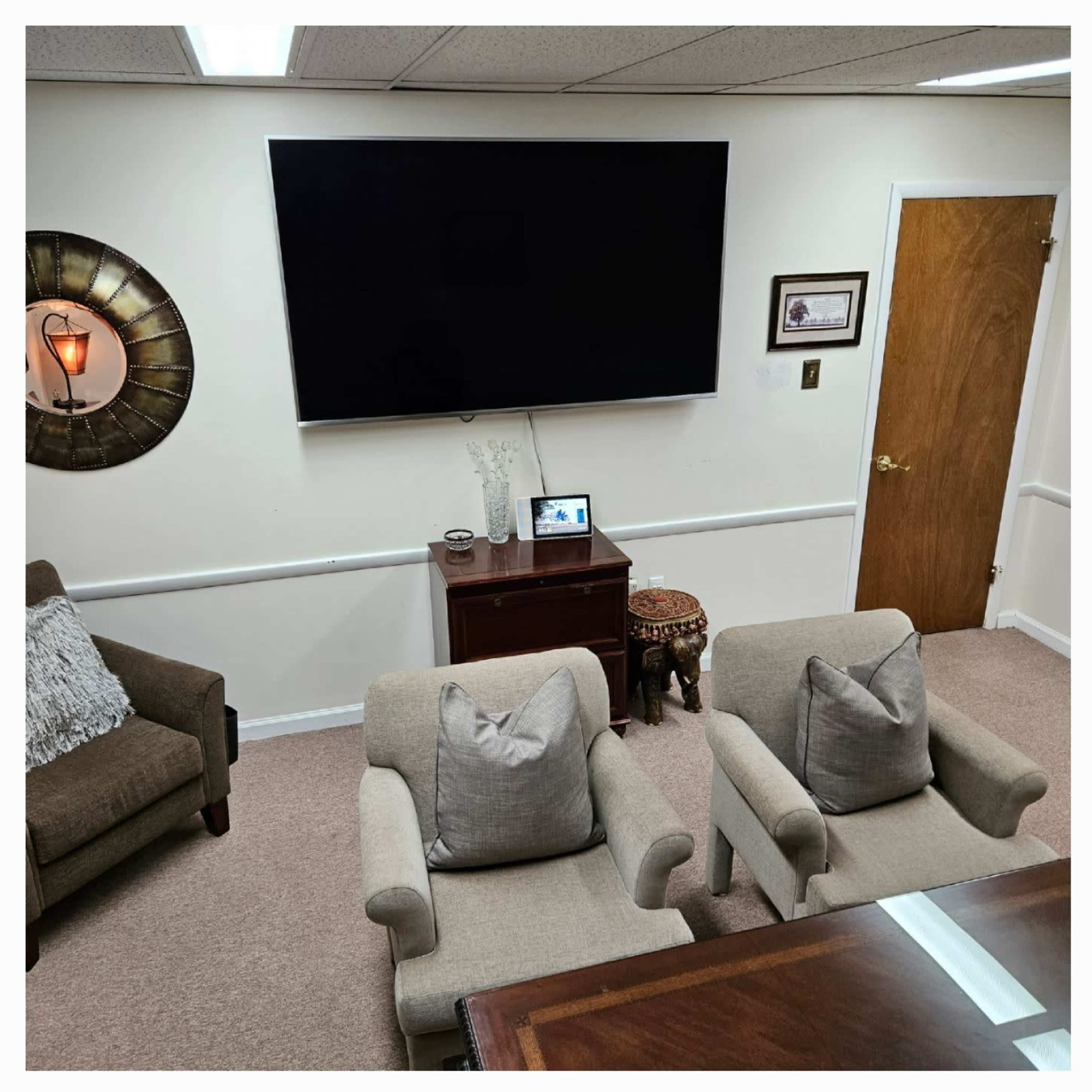 The image shows a tidy office space with two armchairs facing a wooden table, a large television mounted on the wall, and a door leading to another area.