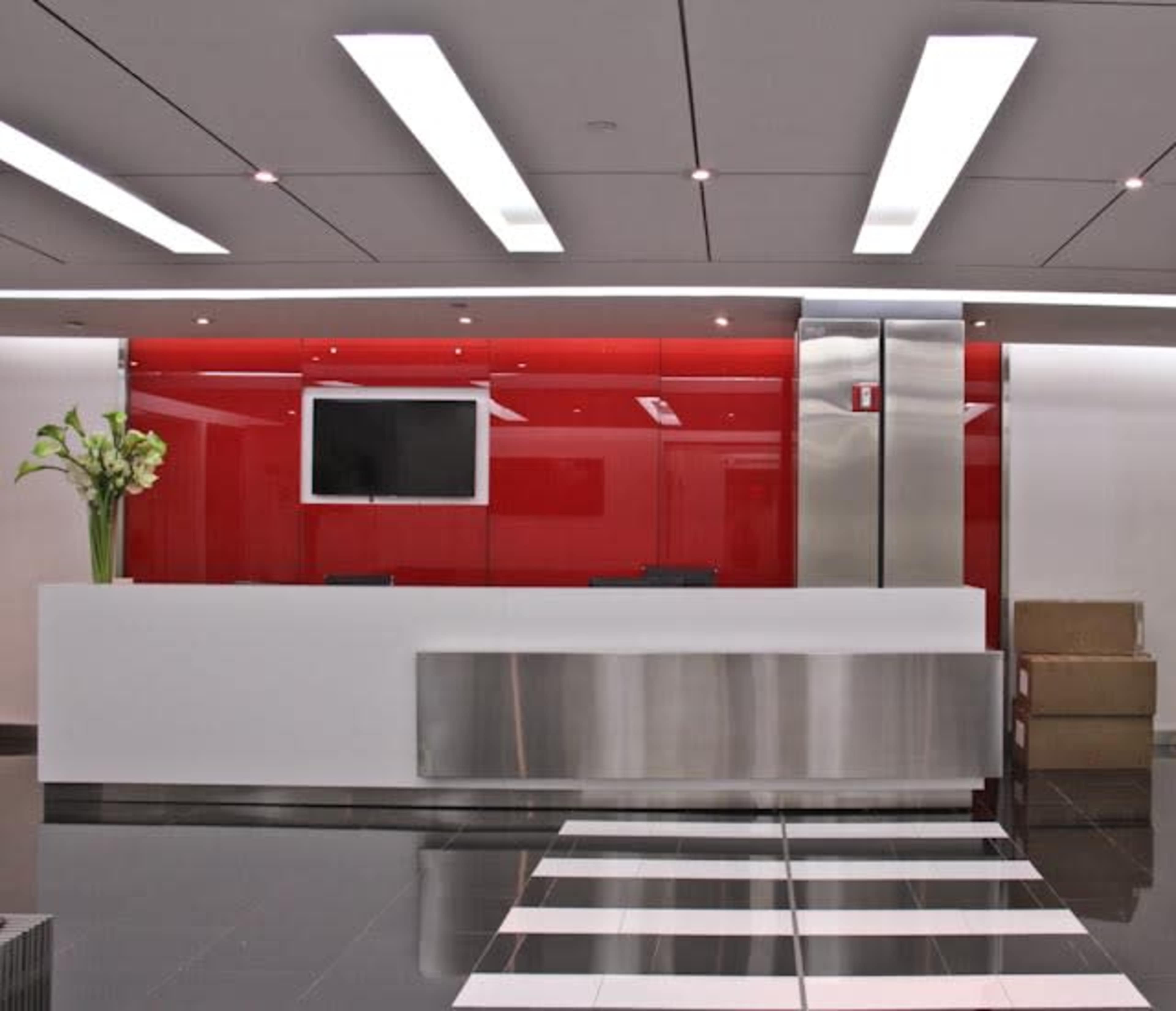 The image shows a modern reception area with a sleek white front desk, red wall panels, and a television mounted on the wall above the desk.