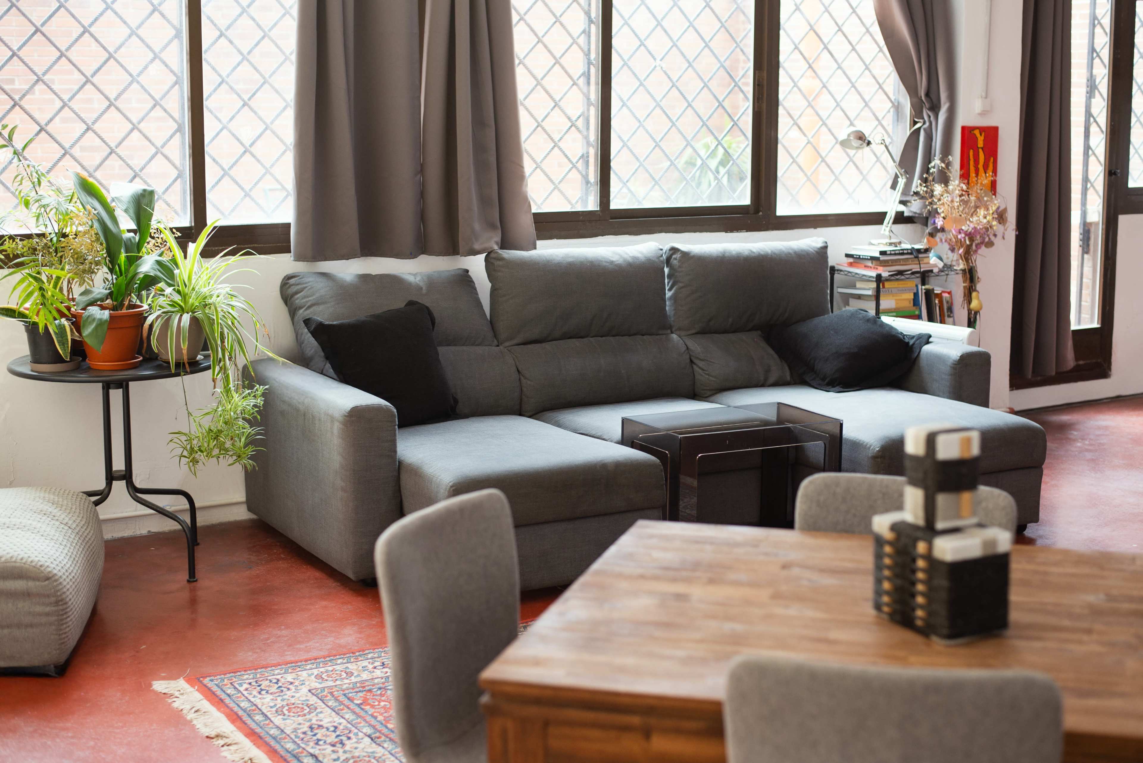 The image shows a cozy living room with a gray sectional sofa, a small black coffee table, and a wooden dining table surrounded by chairs, all set against large windows with greenery outside.