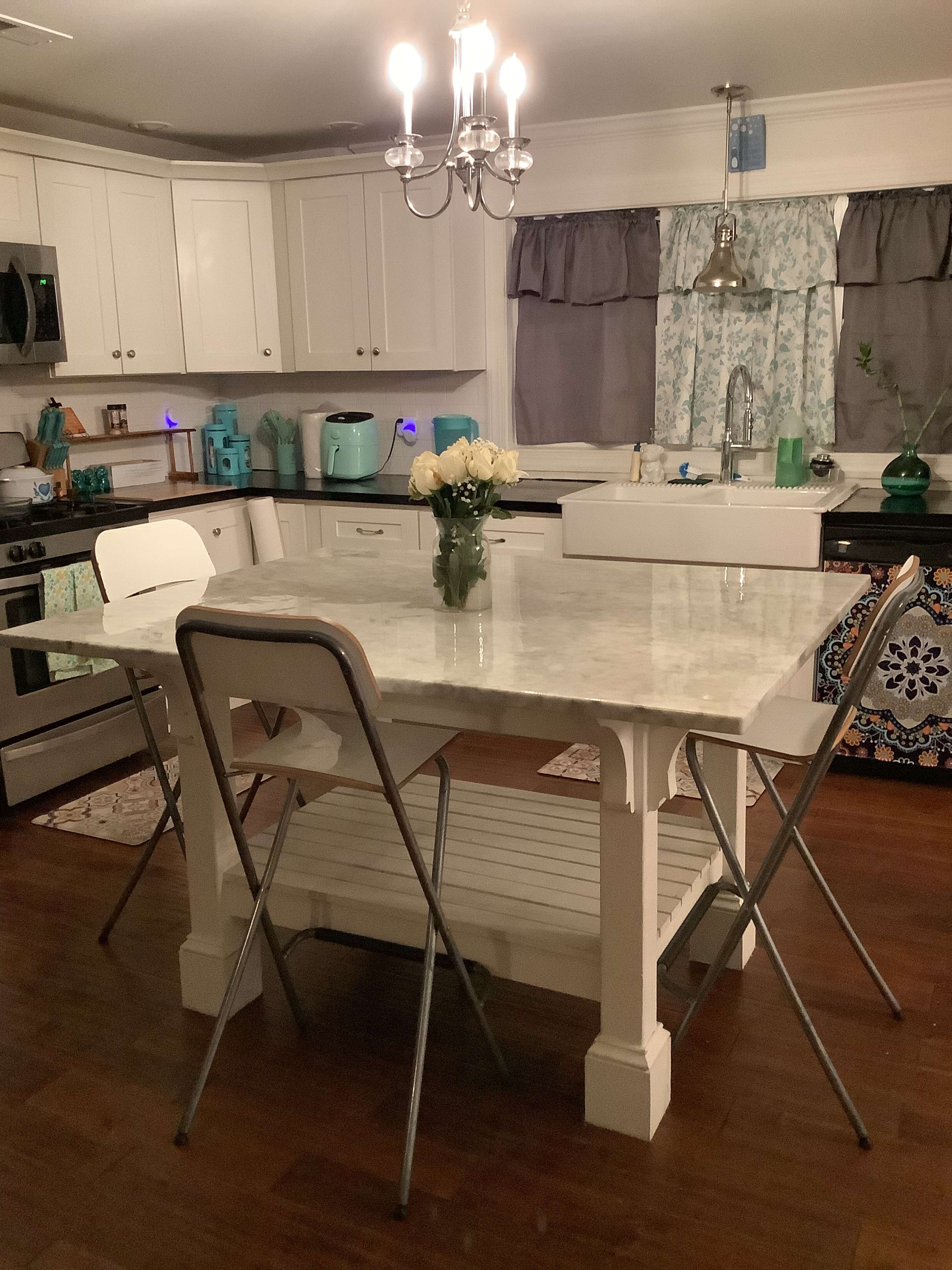 The image shows a modern kitchen with a marble-topped table in the center, surrounded by two folding chairs, and equipped with white cabinets and stainless steel appliances.