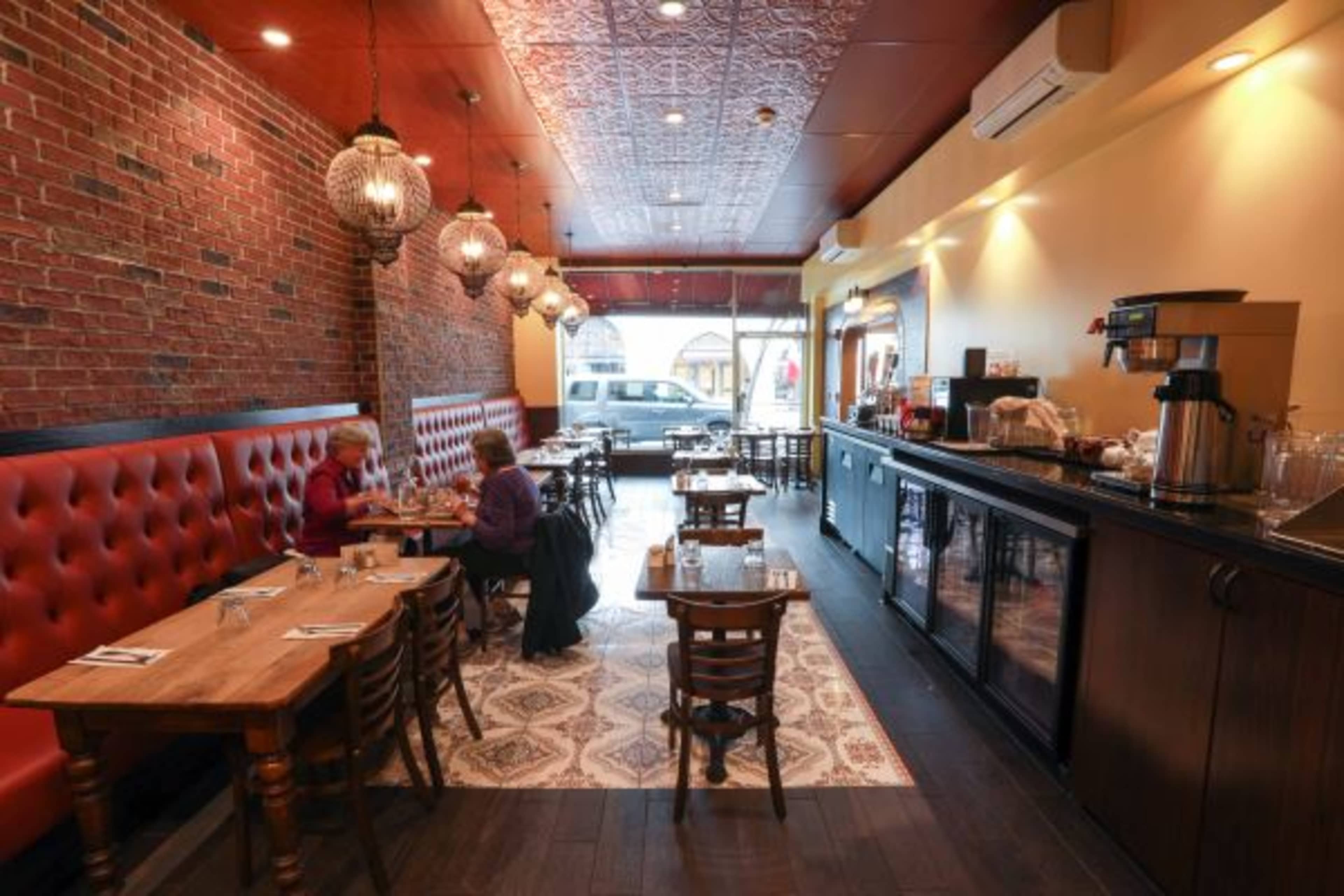 A cozy restaurant interior with wooden tables, red tufted seating, and pendant lights, featuring a brick wall and a serving counter.