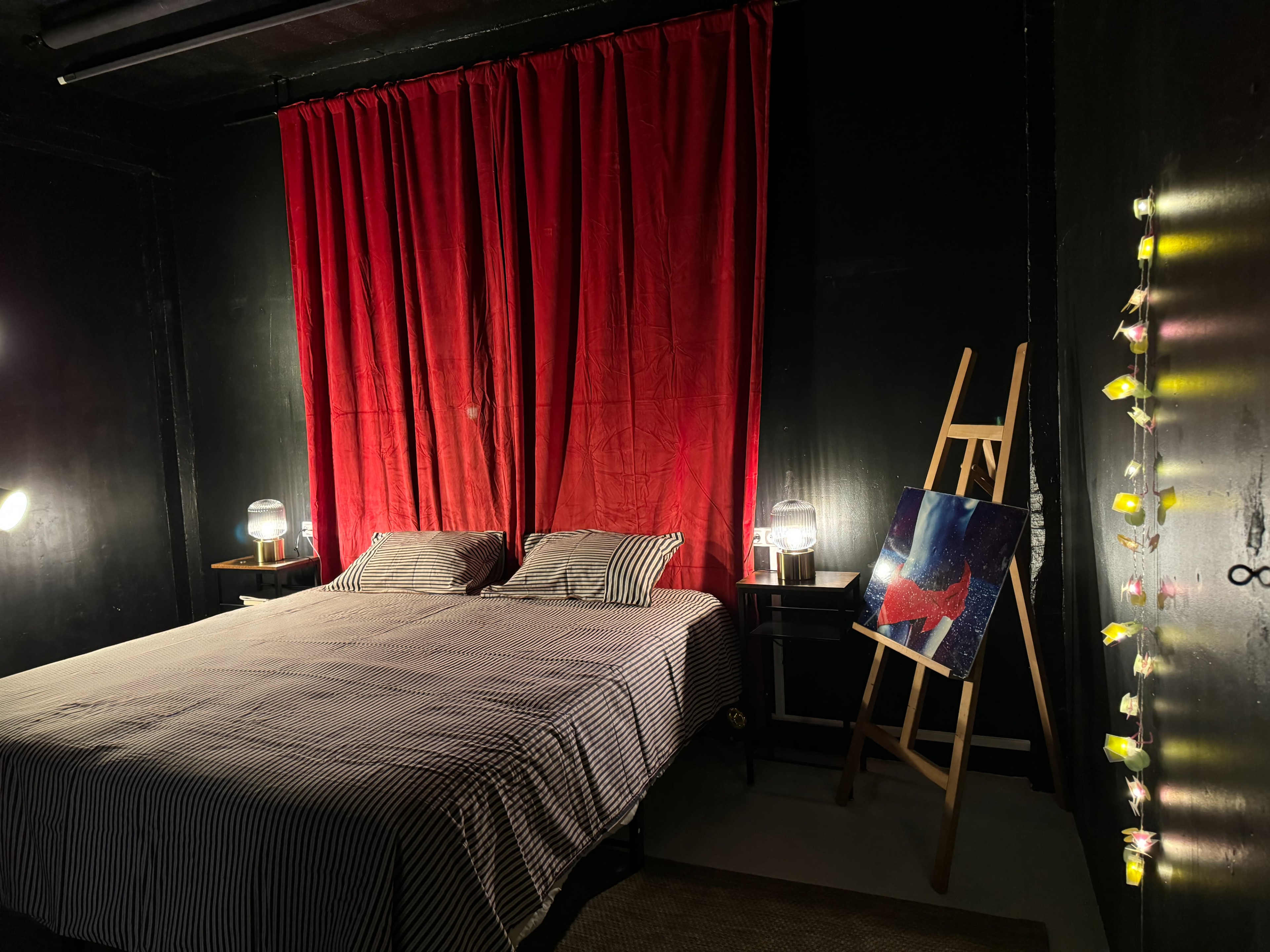 A bedroom with a striped bedspread, red curtains, two bedside lamps, an easel displaying a painting, and string lights on the wall.