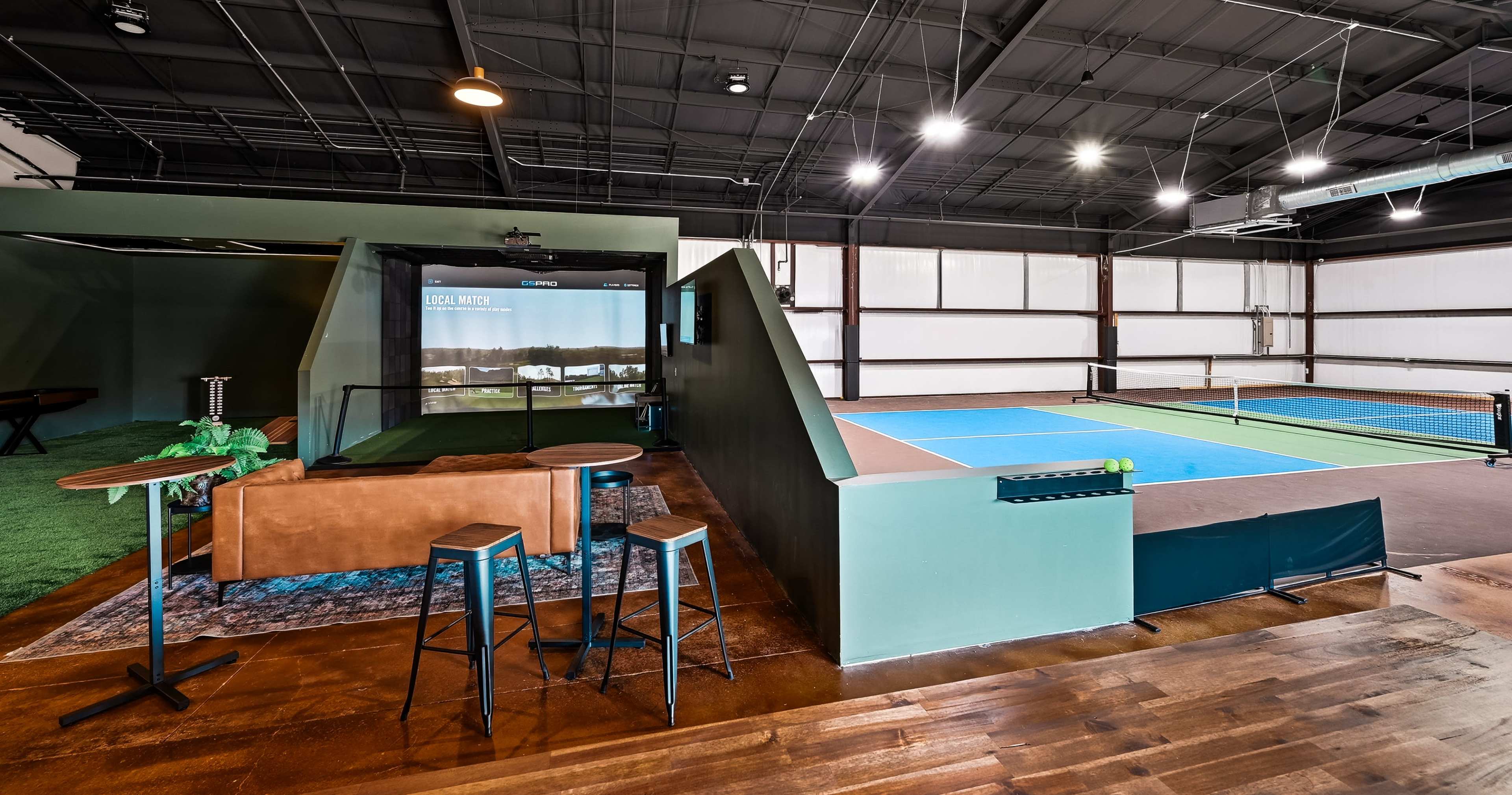 The image shows an indoor sports facility featuring multiple tennis courts, a seating area with bar stools, and a lounge space.