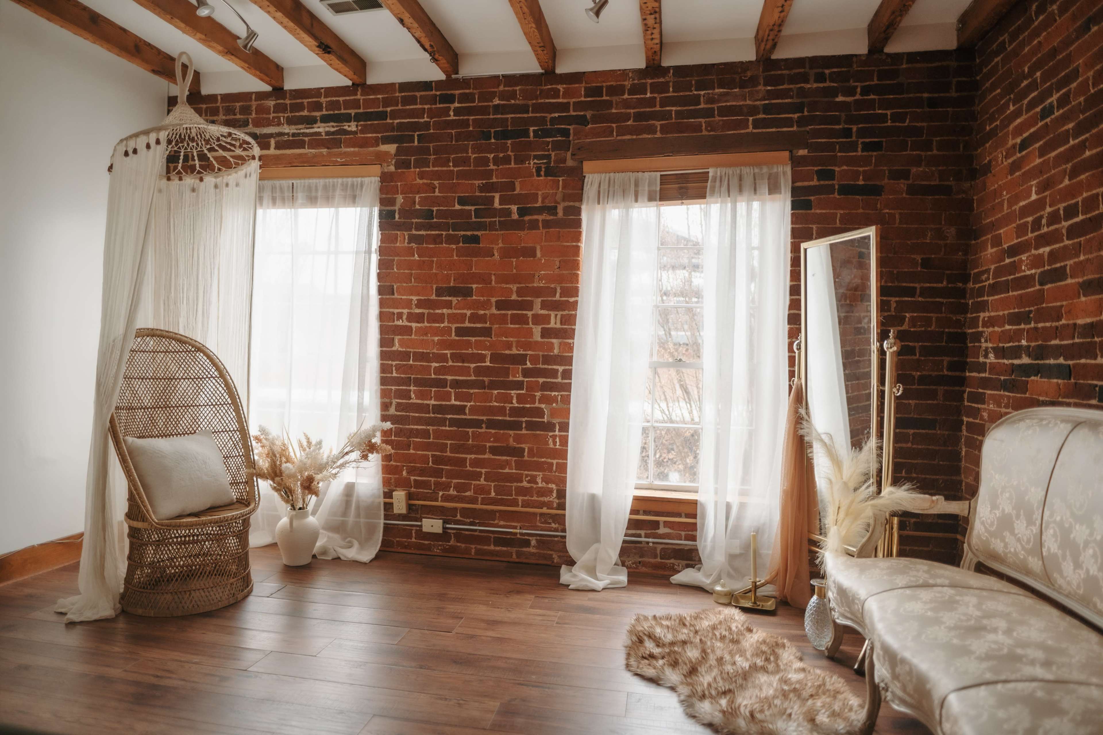 The room features exposed brick walls, two windows draped with sheer curtains, a wicker chair, a decorative mirror, and a small table with dried plants.