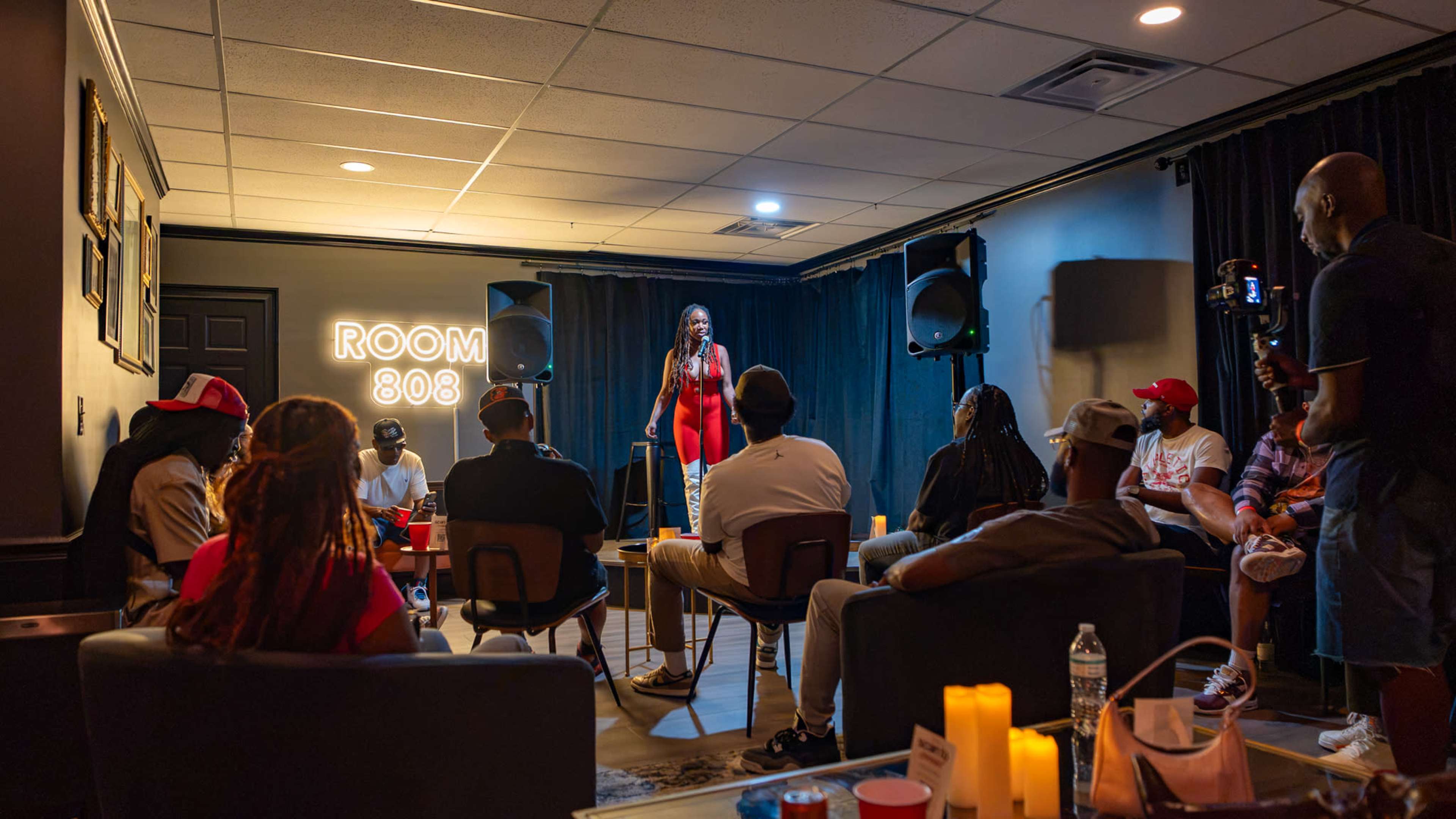 A comedian performs on stage in an intimate room setting while an audience sits in chairs arranged around her.