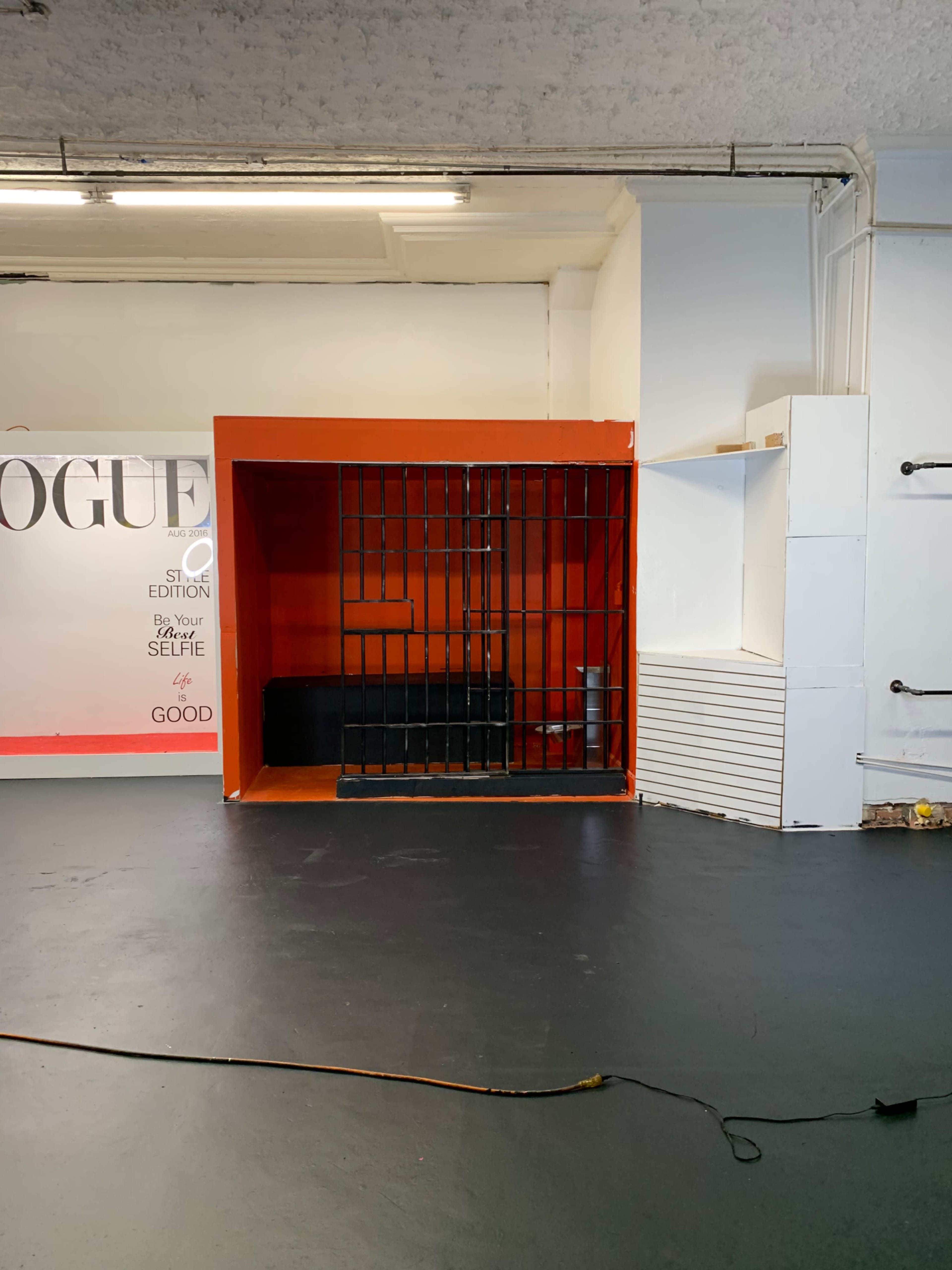 An empty room featuring a red backdrop with a barred enclosure on one side and a white wall with a shelf on the other.