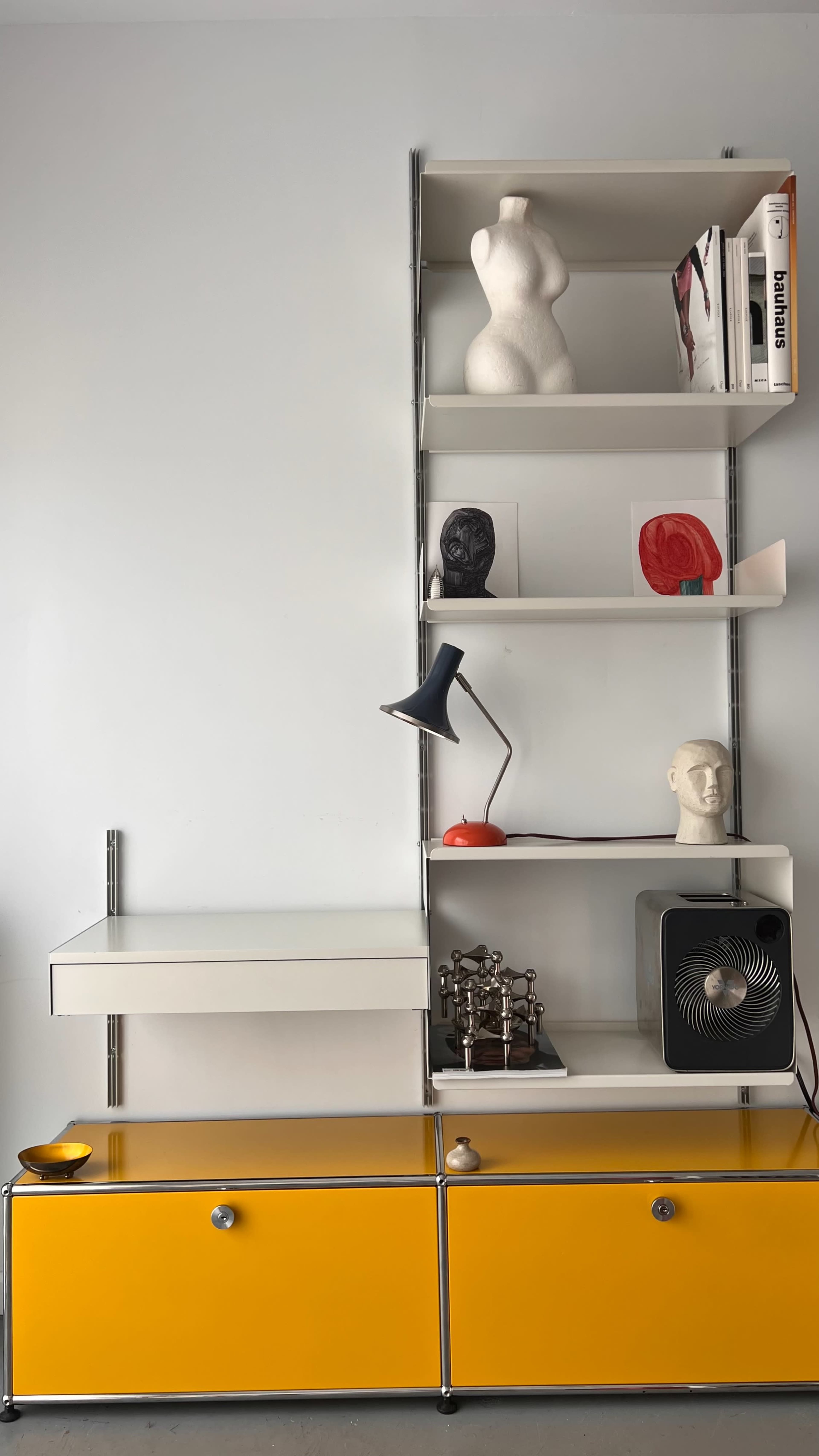 The image features a modern shelving unit against a white wall, displaying various decorative items, books, and a desk with a fan.