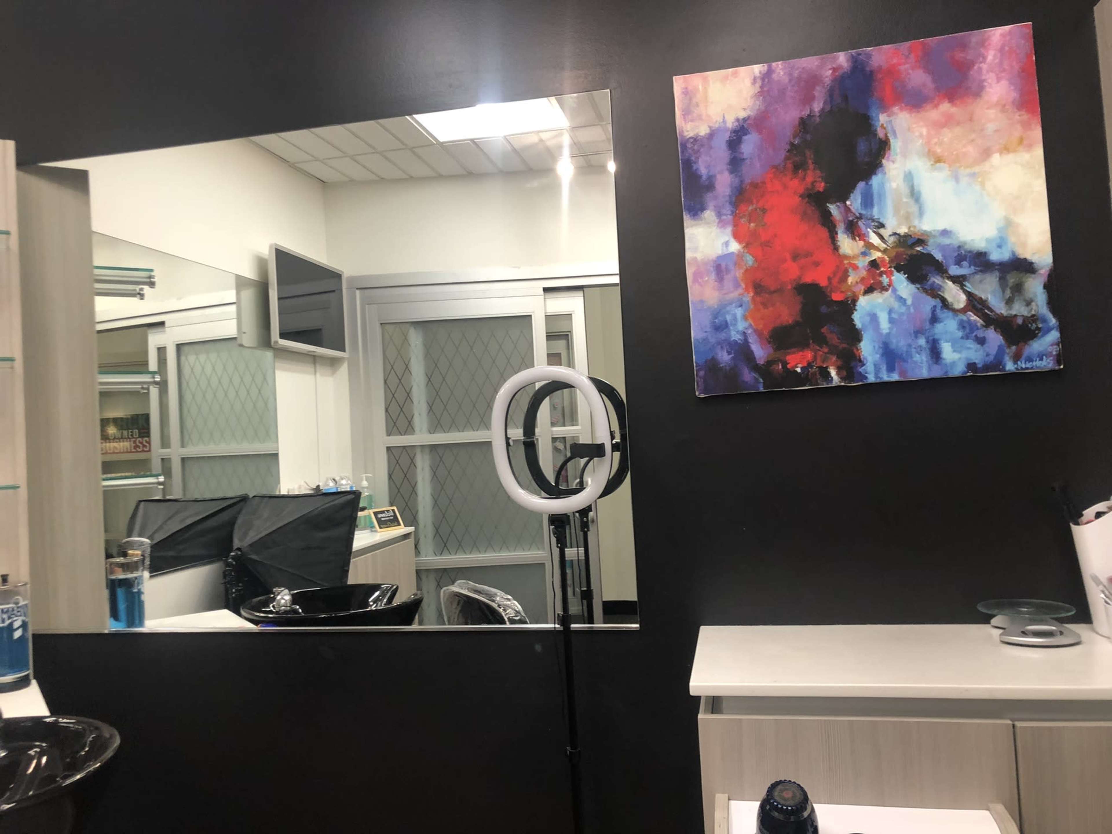 A beauty salon interior featuring a large mirror, a ring light on a stand, and a colorful abstract painting on the wall.