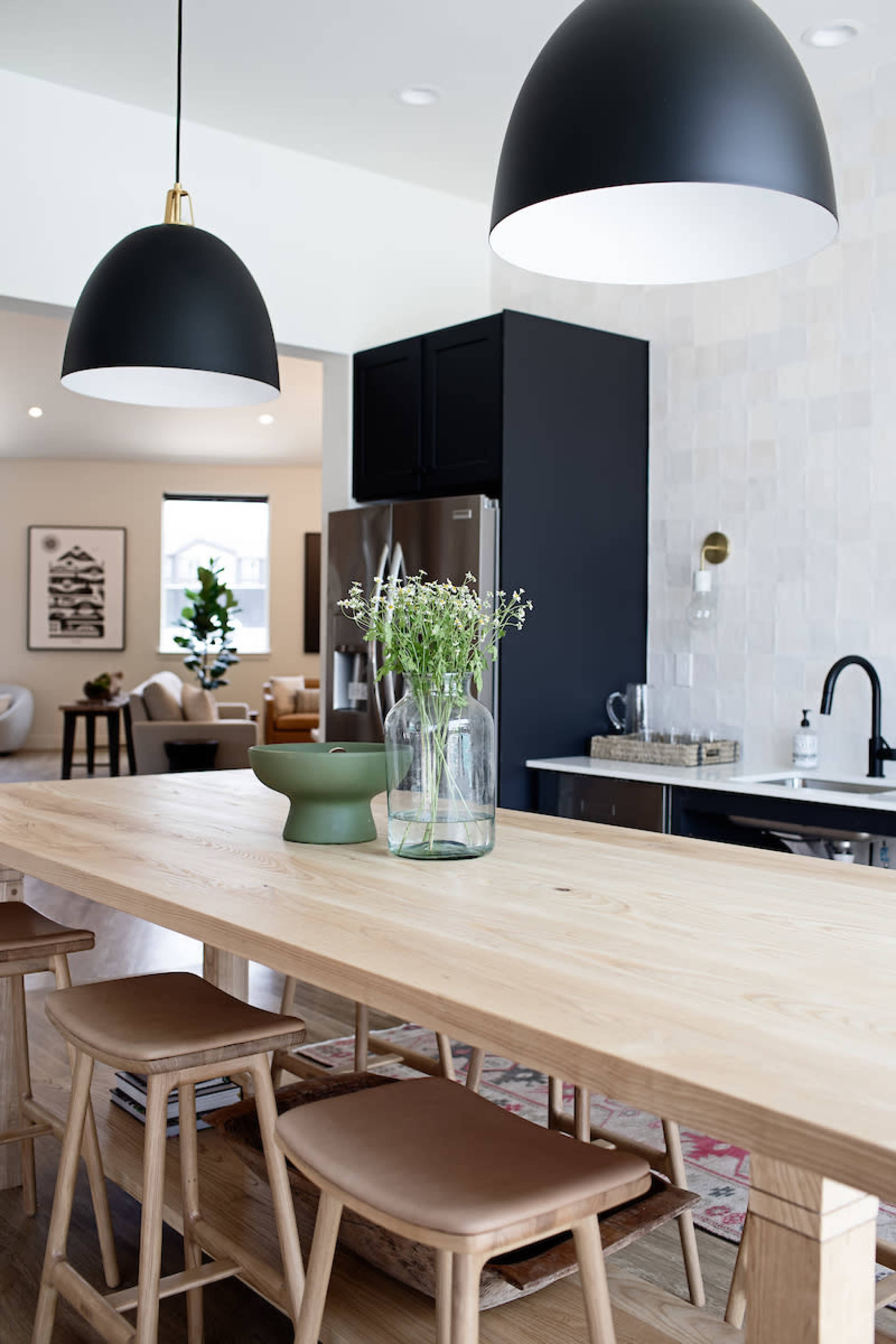 The image features a modern kitchen with a light wooden table, black pendant lights, and a vase of flowers on top, set against a backdrop of stylish cabinetry and a tiled wall.