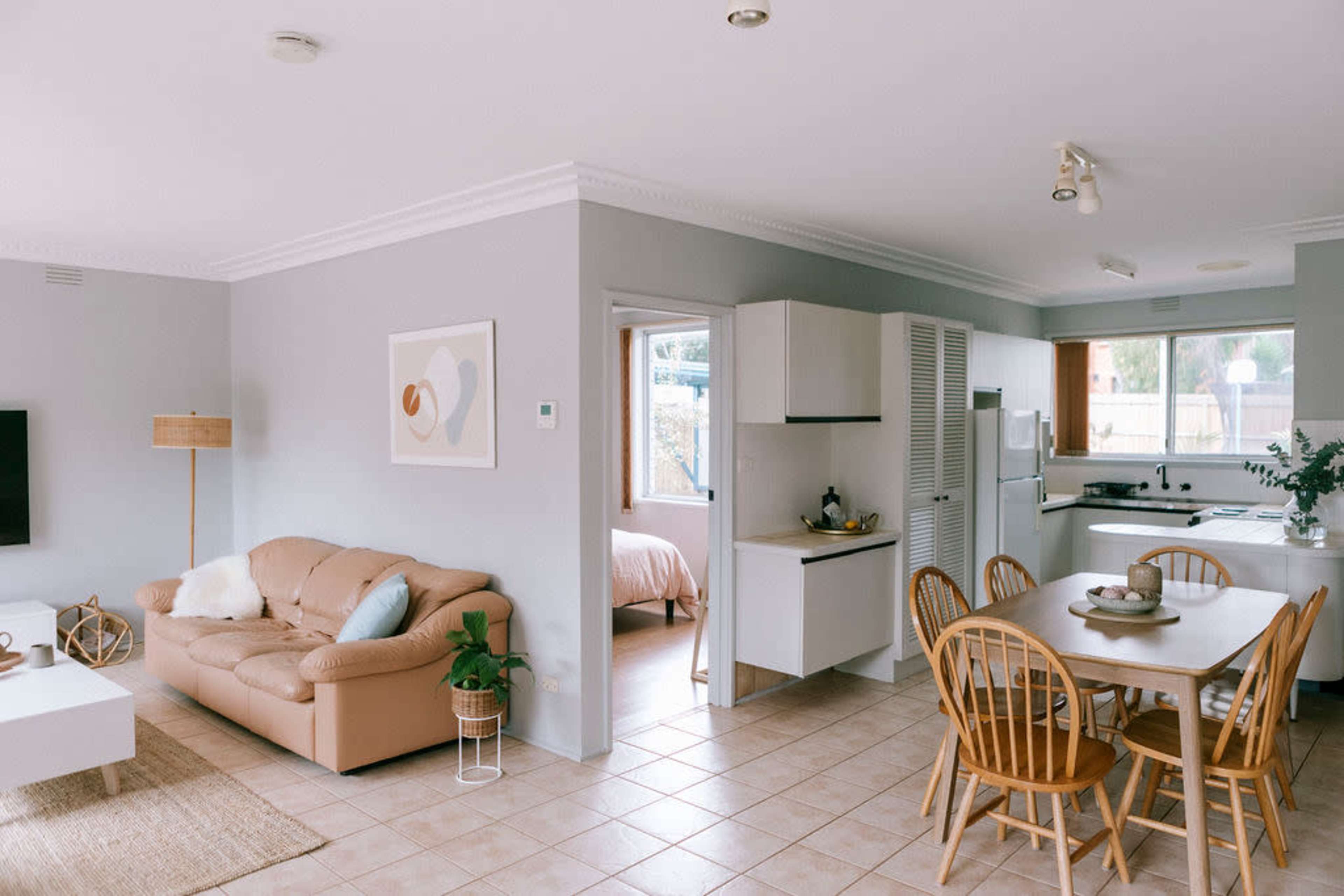 A light-filled living area featuring a beige sofa, a dining table with wooden chairs, and a kitchen visible in the background.