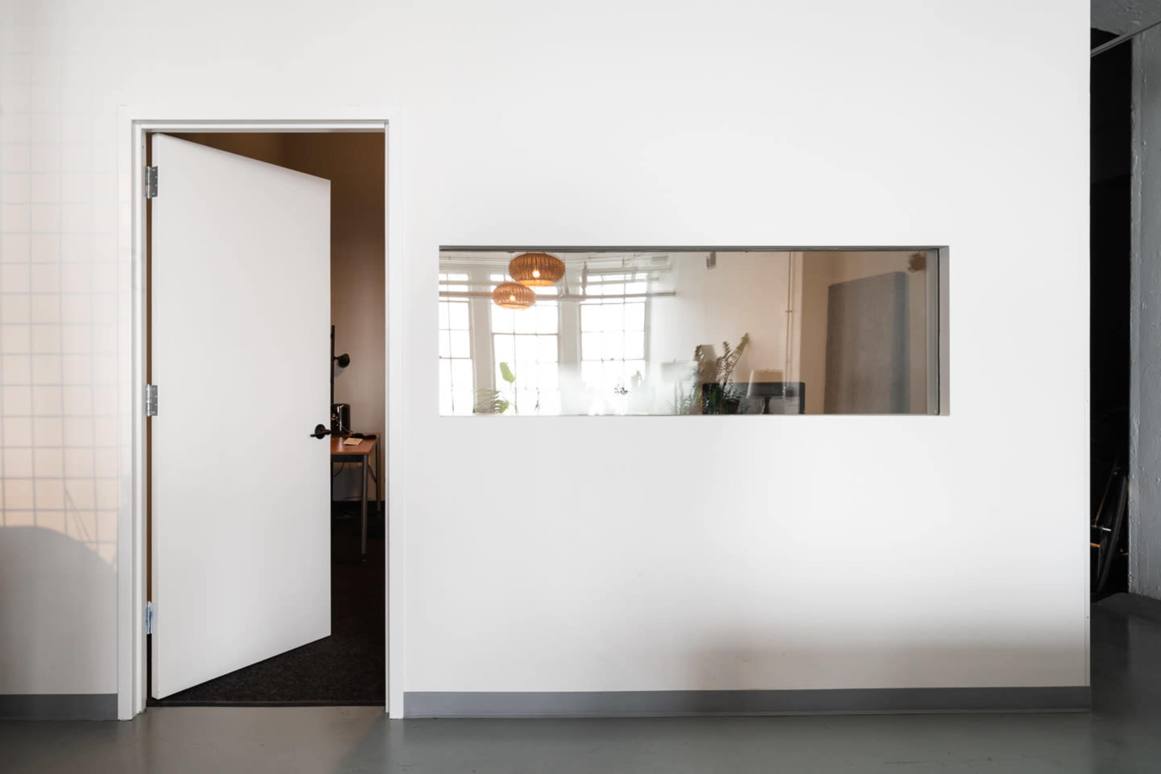 A white wall with a door on the left and a rectangular window revealing a room on the right.
