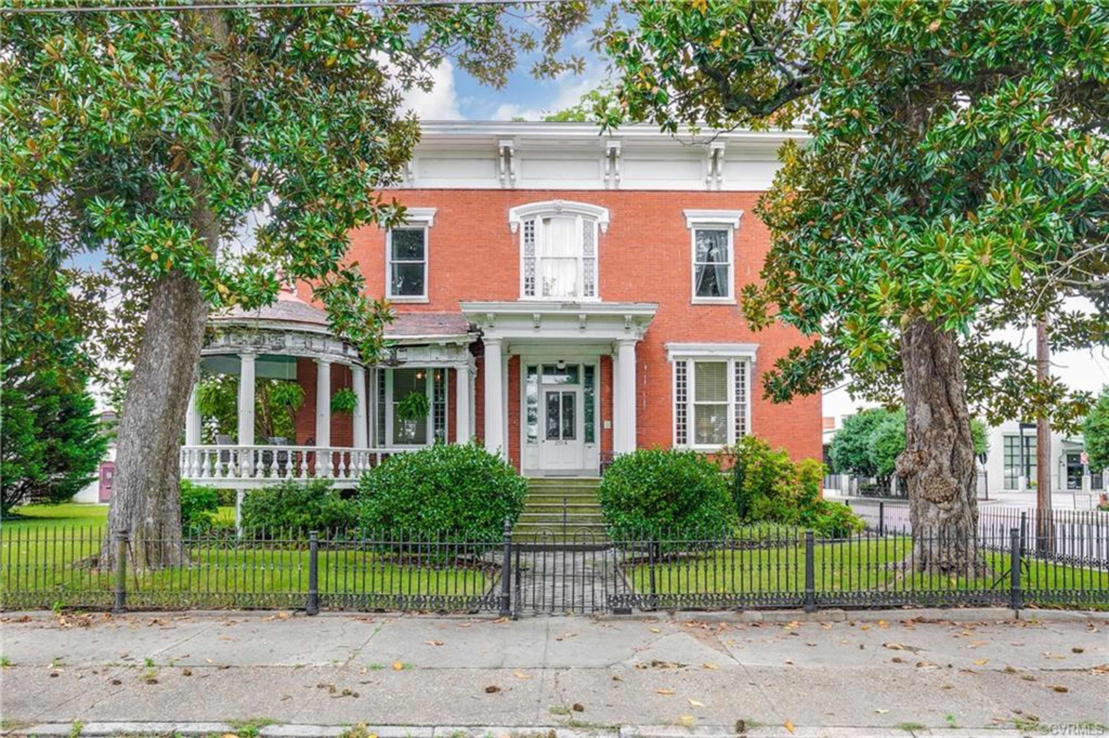 A two-story brick house with a large front porch, surrounded by well-maintained lawns and trees, enclosed by an iron fence.