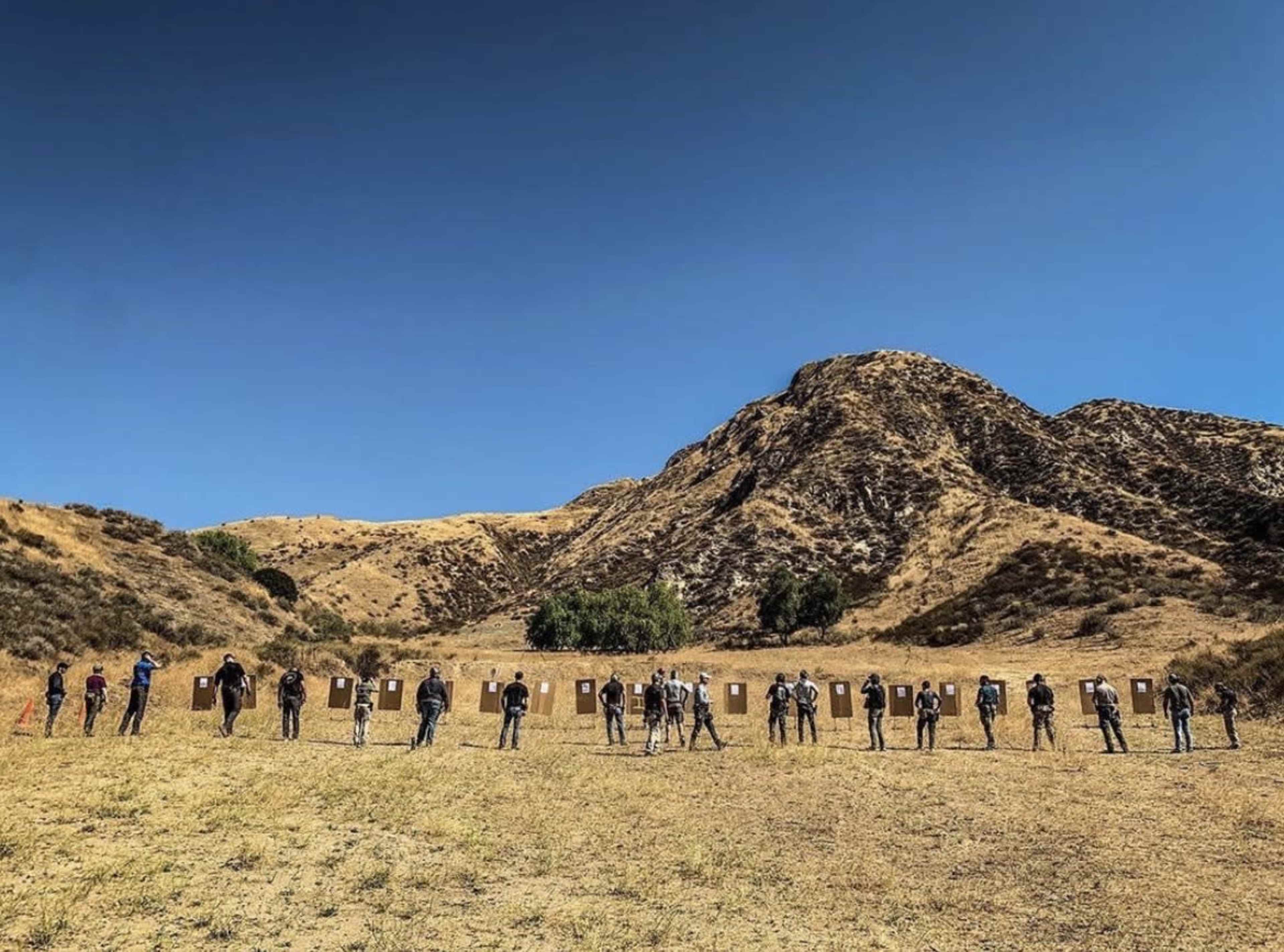 A group of individuals stands in a line, aiming at target boards set up on a hillside under a clear blue sky.