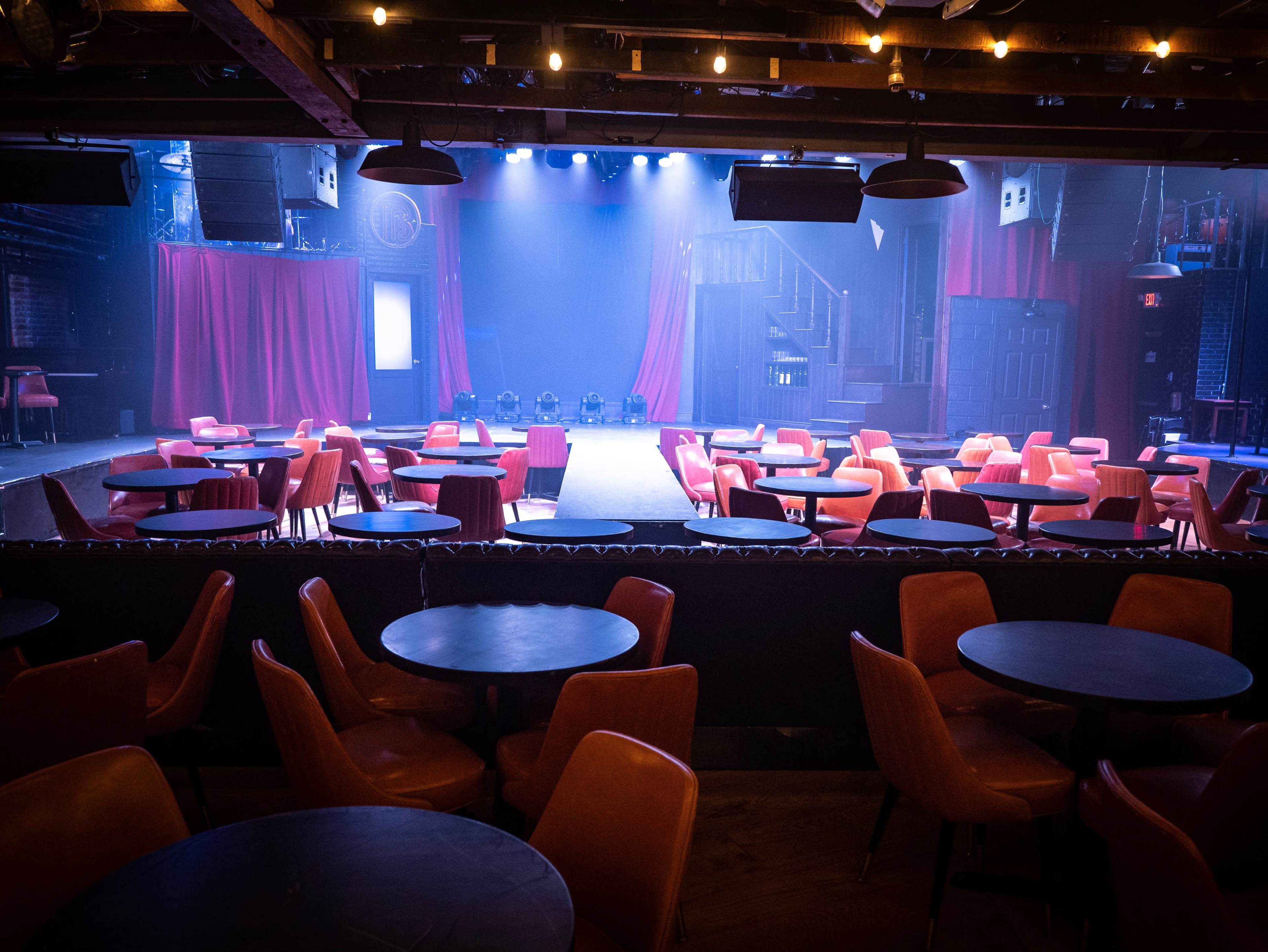 An empty performance venue with round tables and chairs arranged in front of a dimly lit stage framed by red curtains.