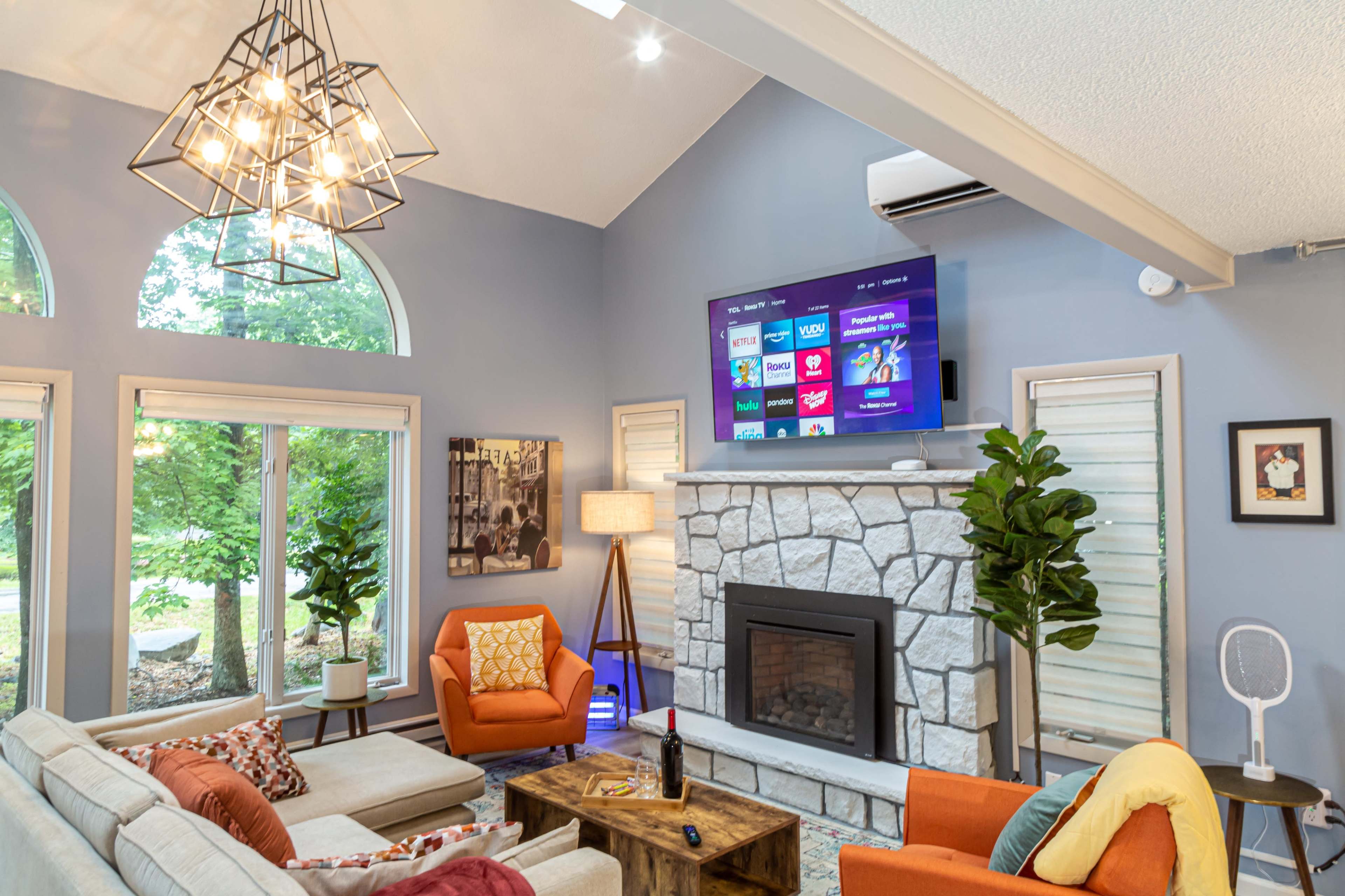 A modern living room features a stone fireplace, a large wall-mounted TV displaying streaming options, and comfortable seating arranged around a wooden coffee table.