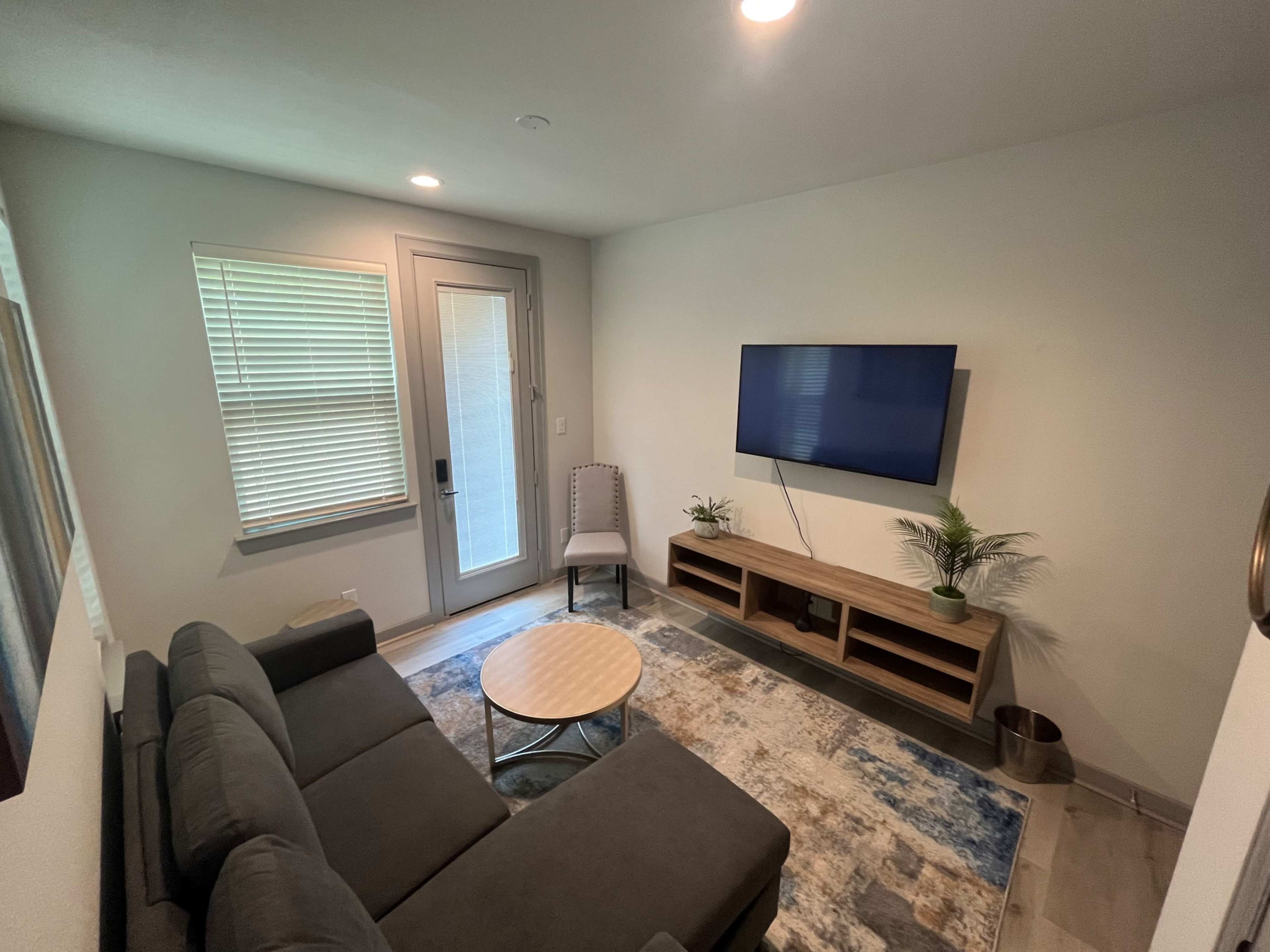 The room features a grey sectional sofa, a round coffee table, a television mounted on the wall, and a door leading outside, with a small chair nearby.