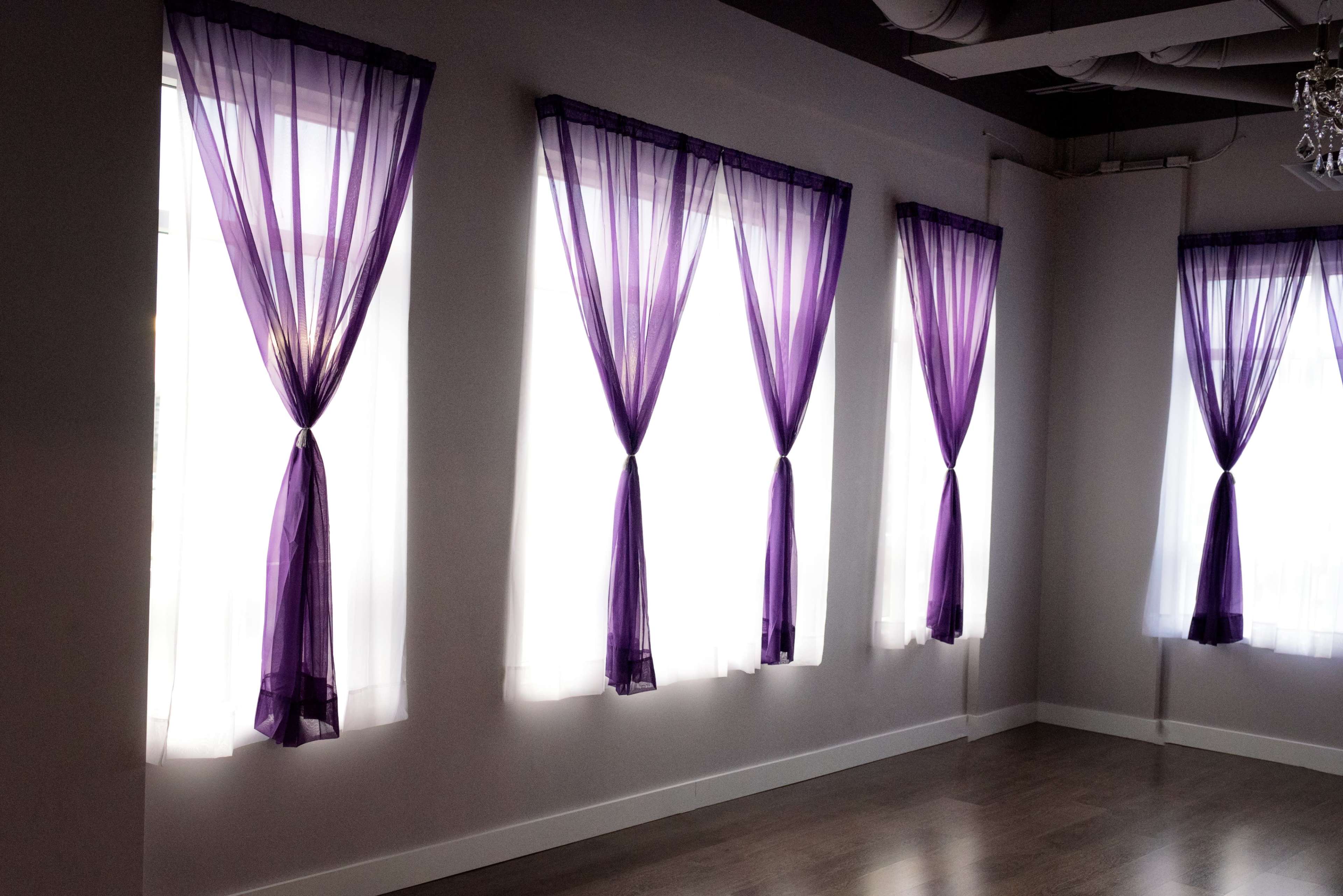 Four windows draped with sheer purple curtains in a well-lit room with wooden flooring.