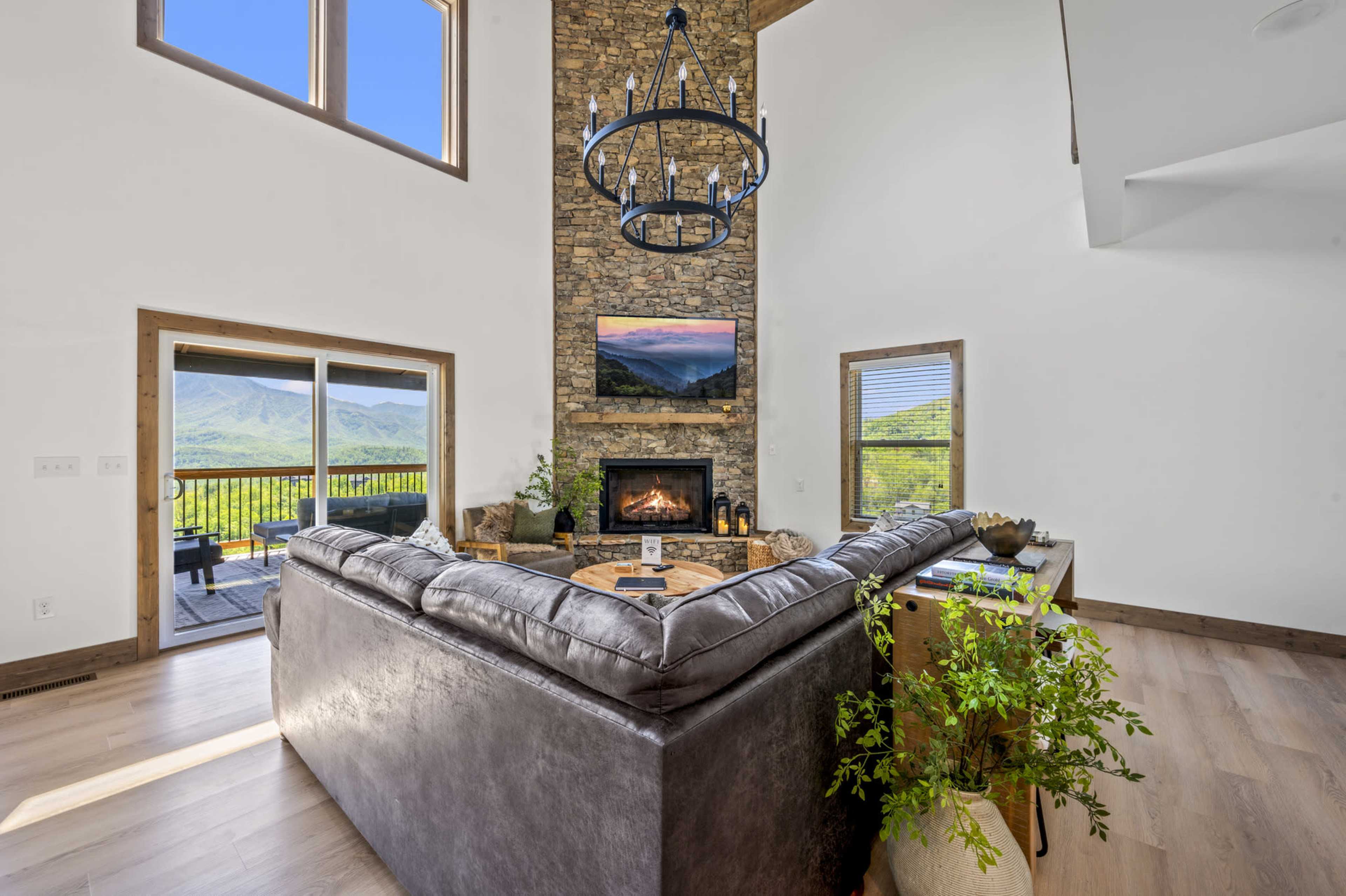 A modern living room with a large sectional sofa facing a stone fireplace, a mounted television above it, and large windows offering a view of the mountains.