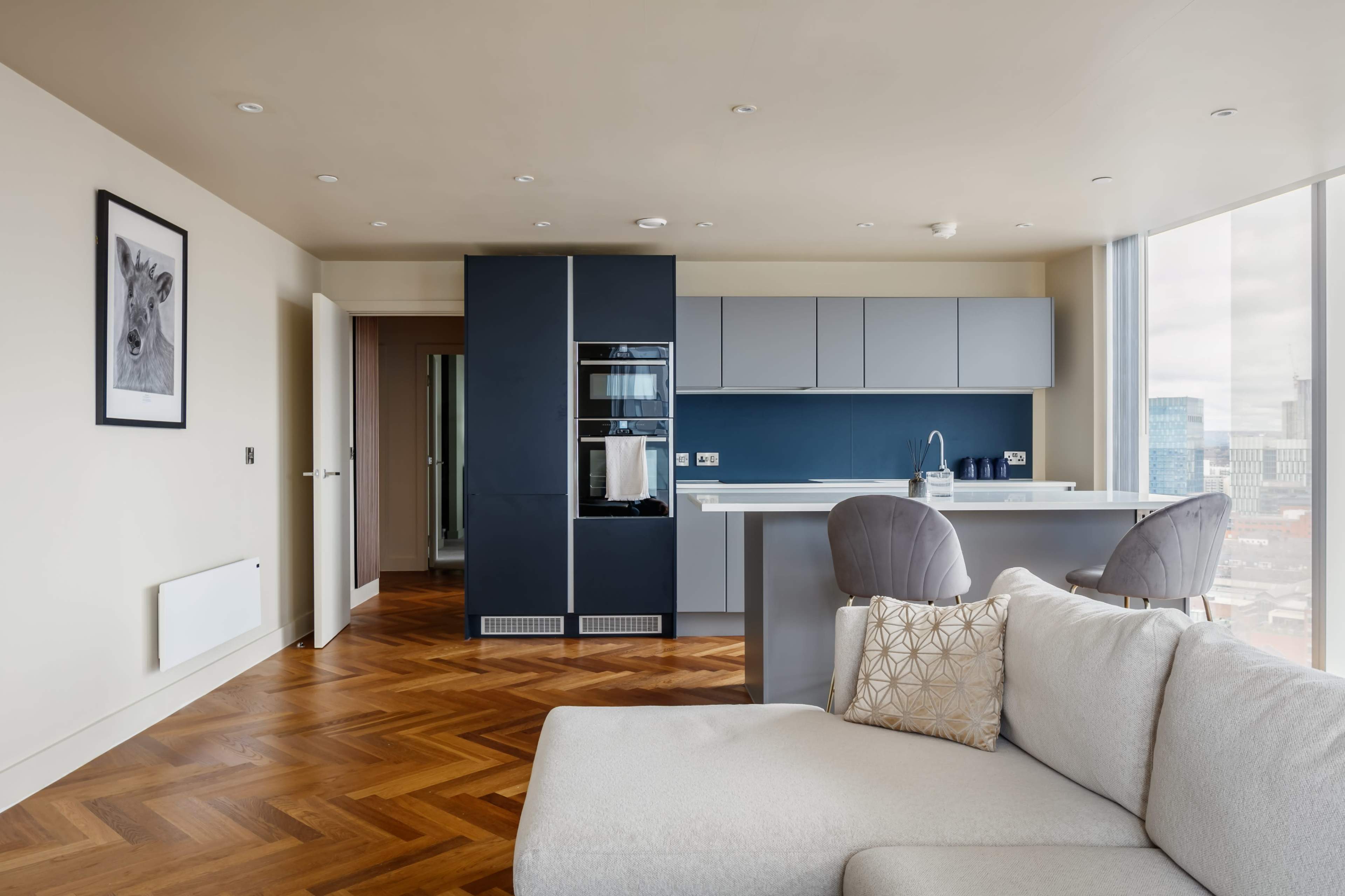 A modern kitchen and living area features a sleek design with a blue kitchen cabinet unit, hardwood flooring, and large windows providing a view of the city.