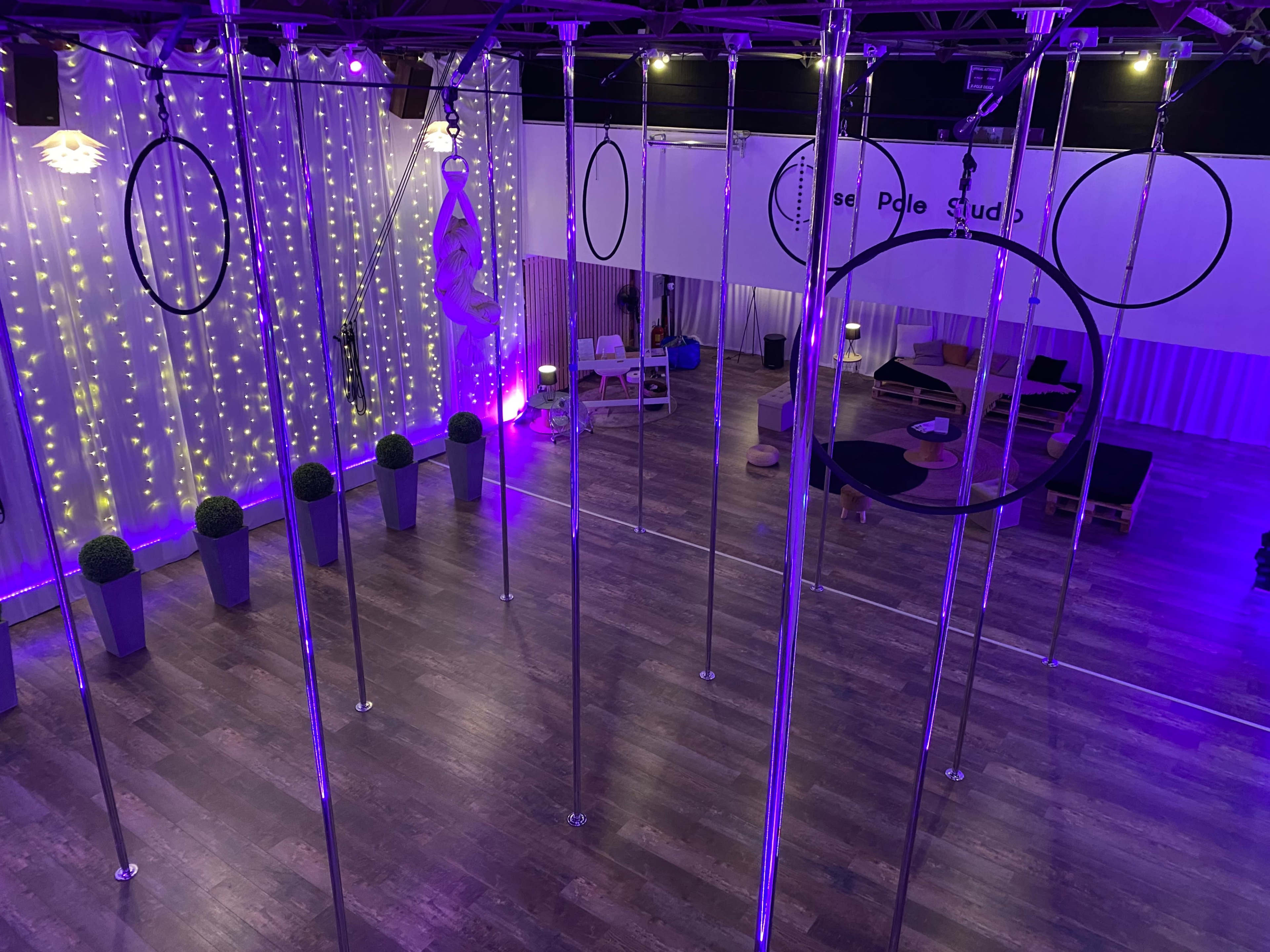 An aerial studio with several poles and hoops, illuminated by purple lights and decorated with string lights in the background.