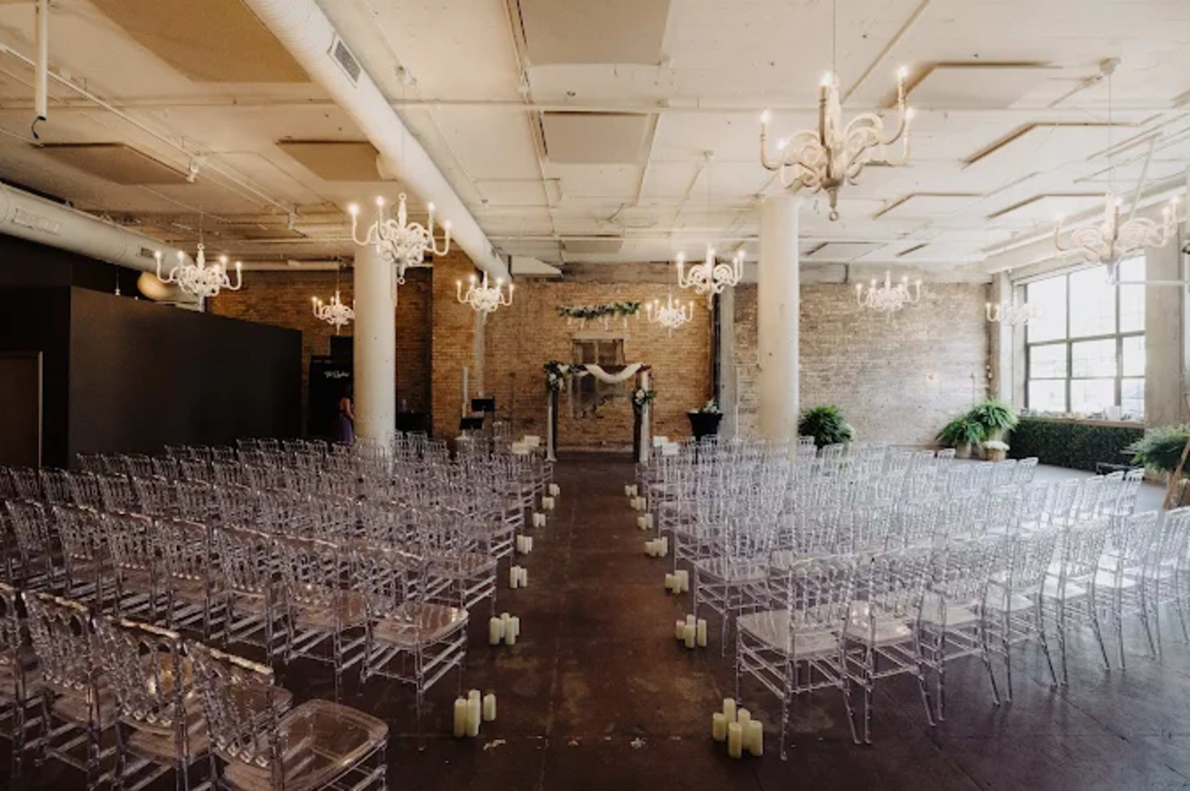 The image depicts a spacious wedding ceremony setup with clear chairs arranged in rows facing a decorated altar, illuminated by chandeliers and surrounded by greenery.