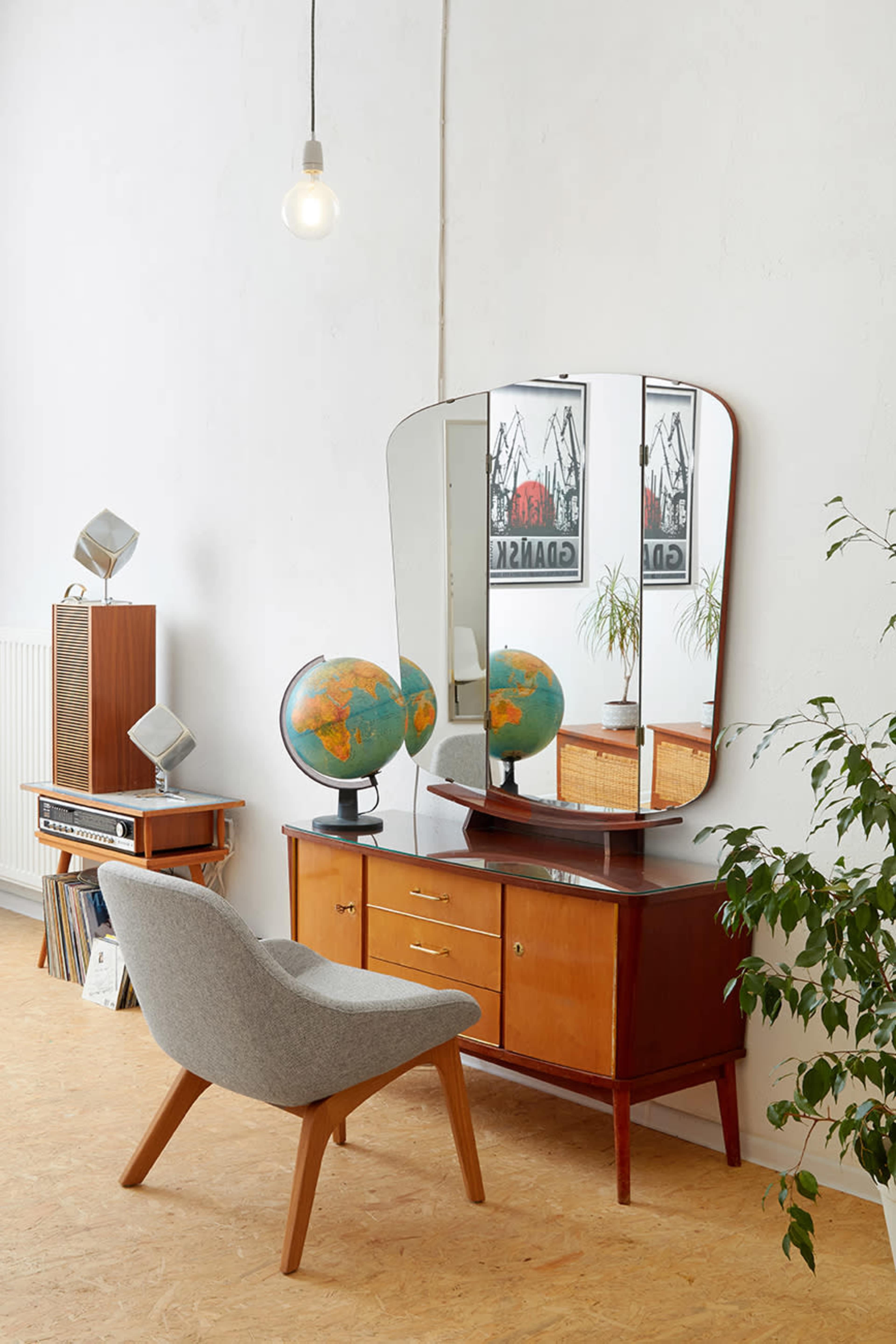 A mid-century modern dresser with a large mirror is positioned against a white wall, accompanied by two globes, a plant, and a retro speaker.