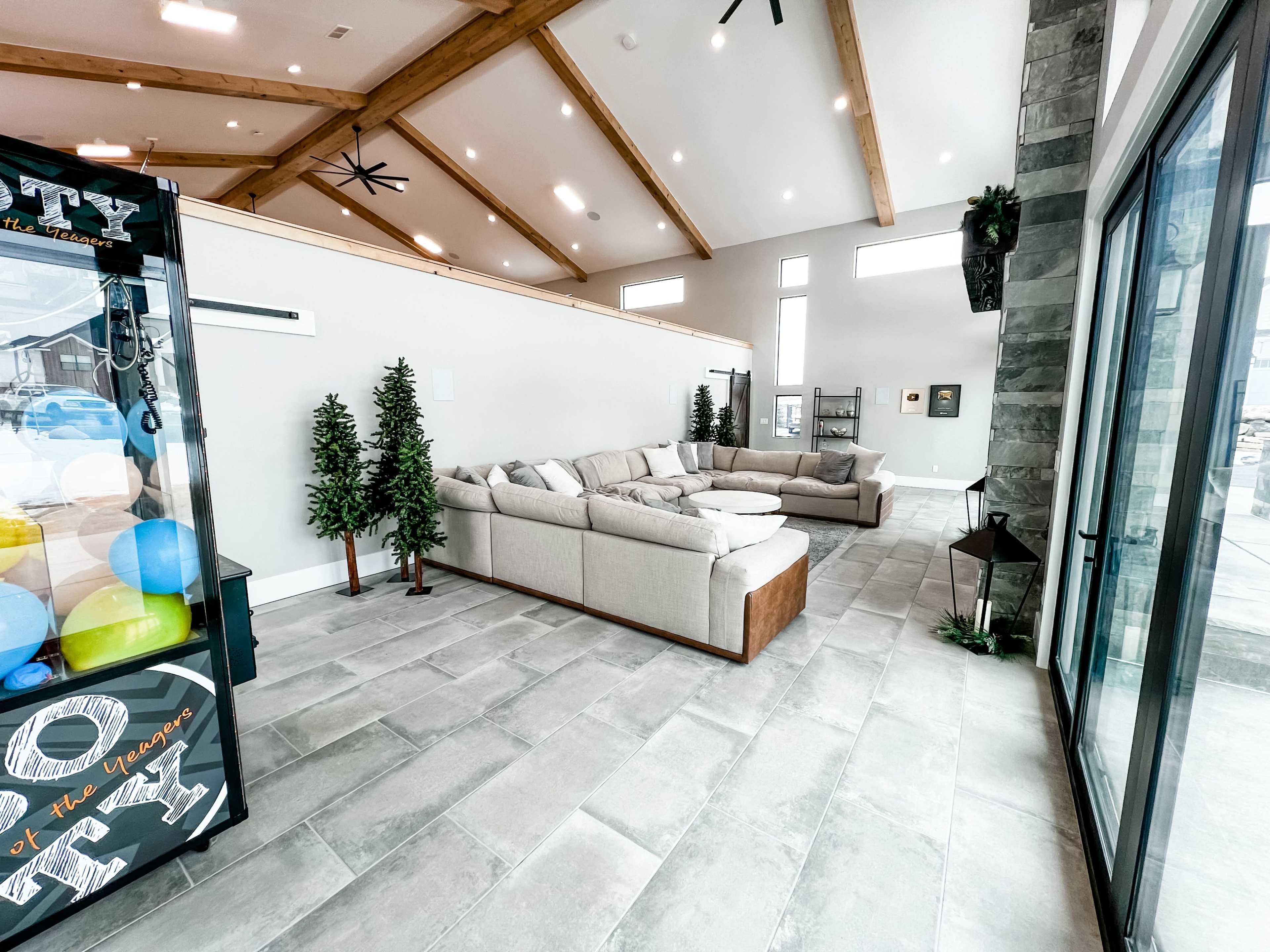 A spacious living area featuring a light-colored sectional sofa, potted artificial trees, and large windows, with a game arcade machine in one corner.