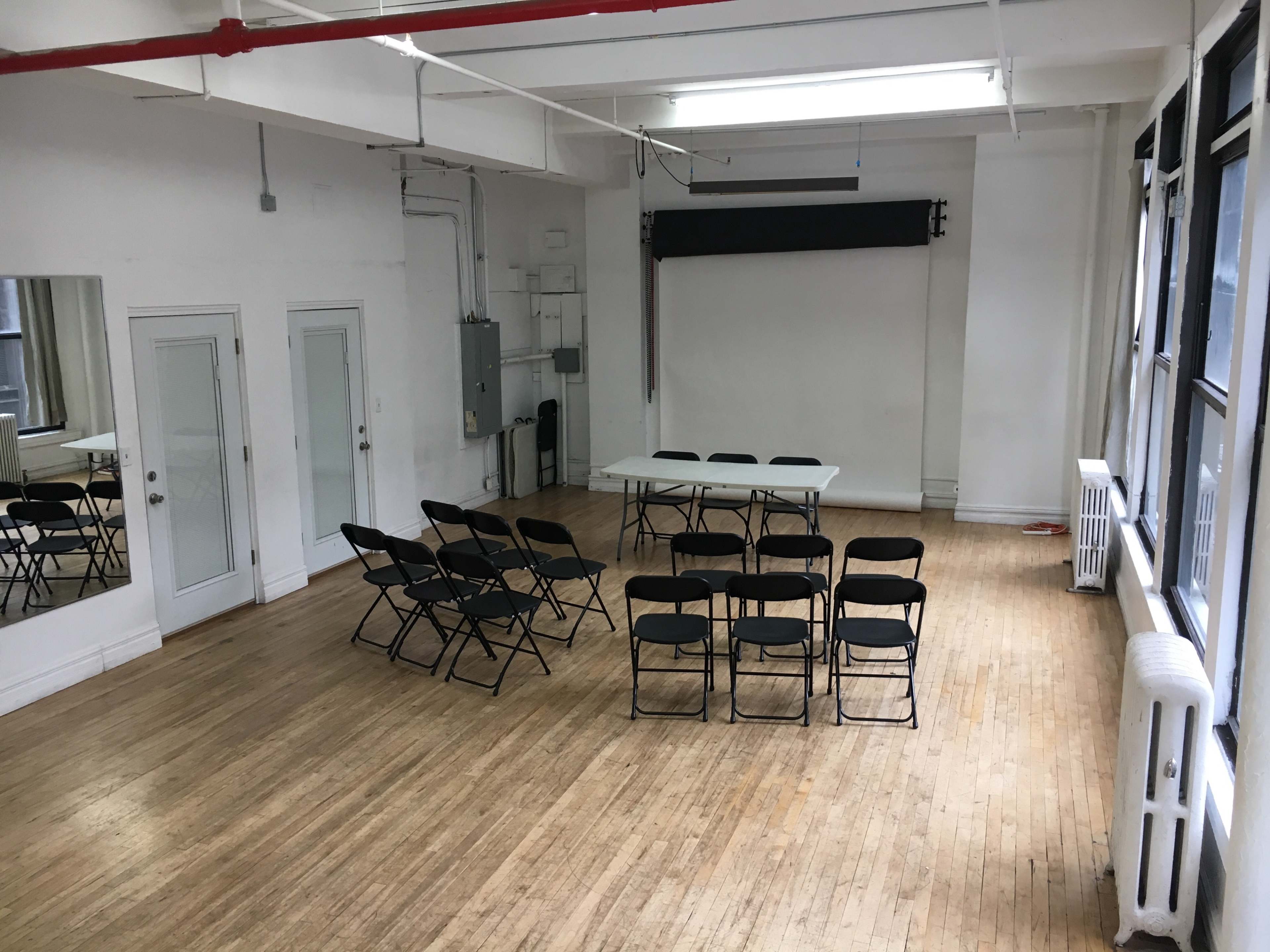 A spacious room with wooden floors containing a table surrounded by black folding chairs and large windows.