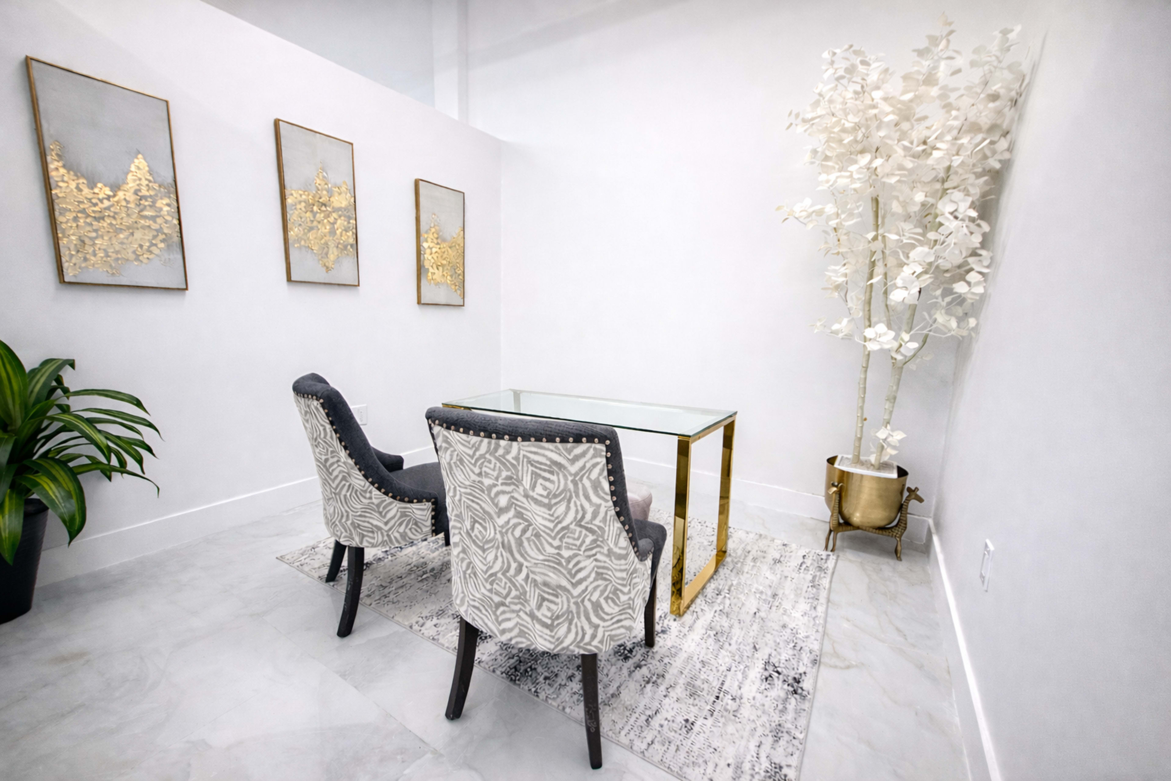 The image shows a modern minimalistic office space featuring a glass-top desk, two upholstered chairs, and a decorative tree in a metallic planter.