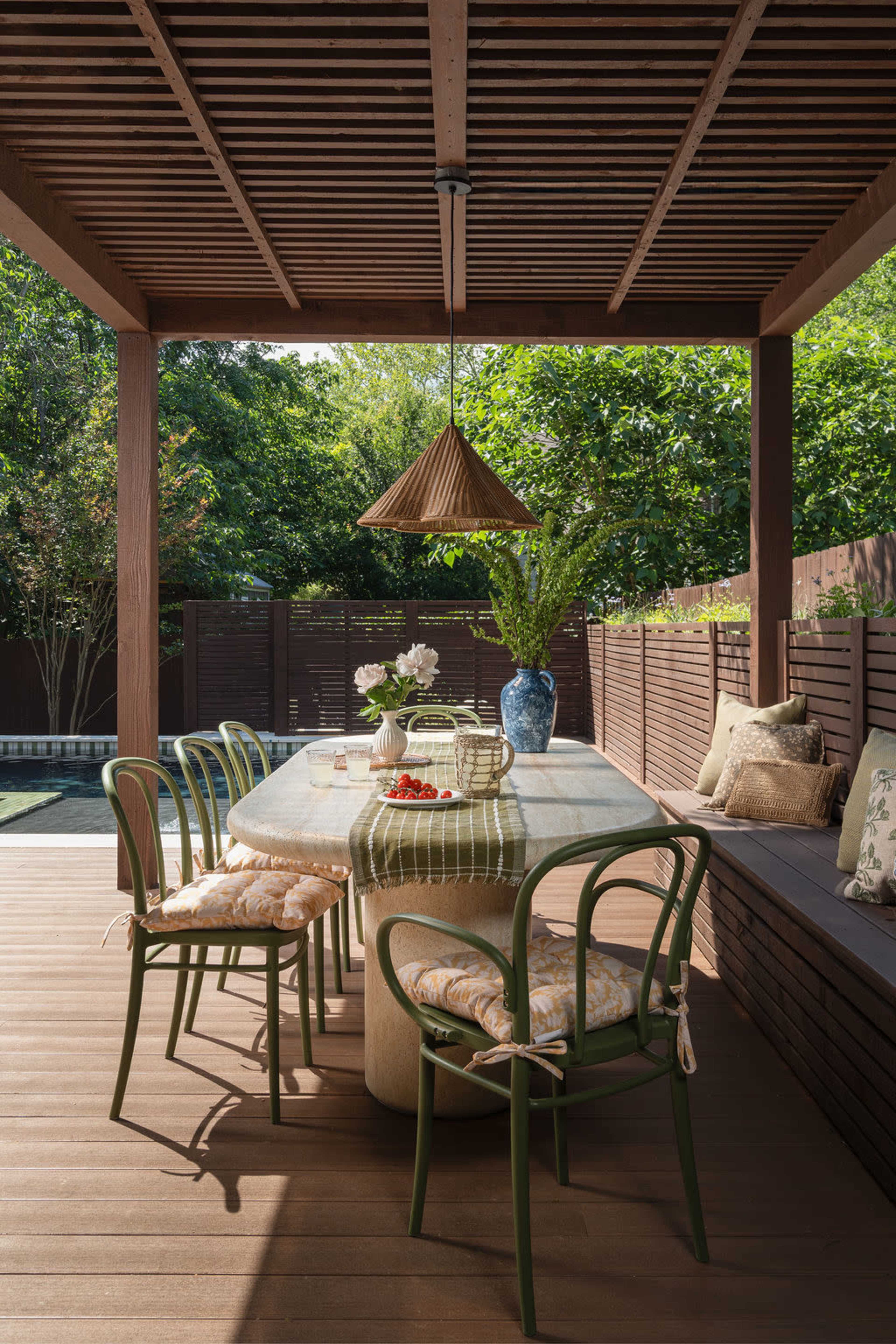 An outdoor dining area features a long table surrounded by green chairs, with a pendant light hanging above and lush greenery in the background.
