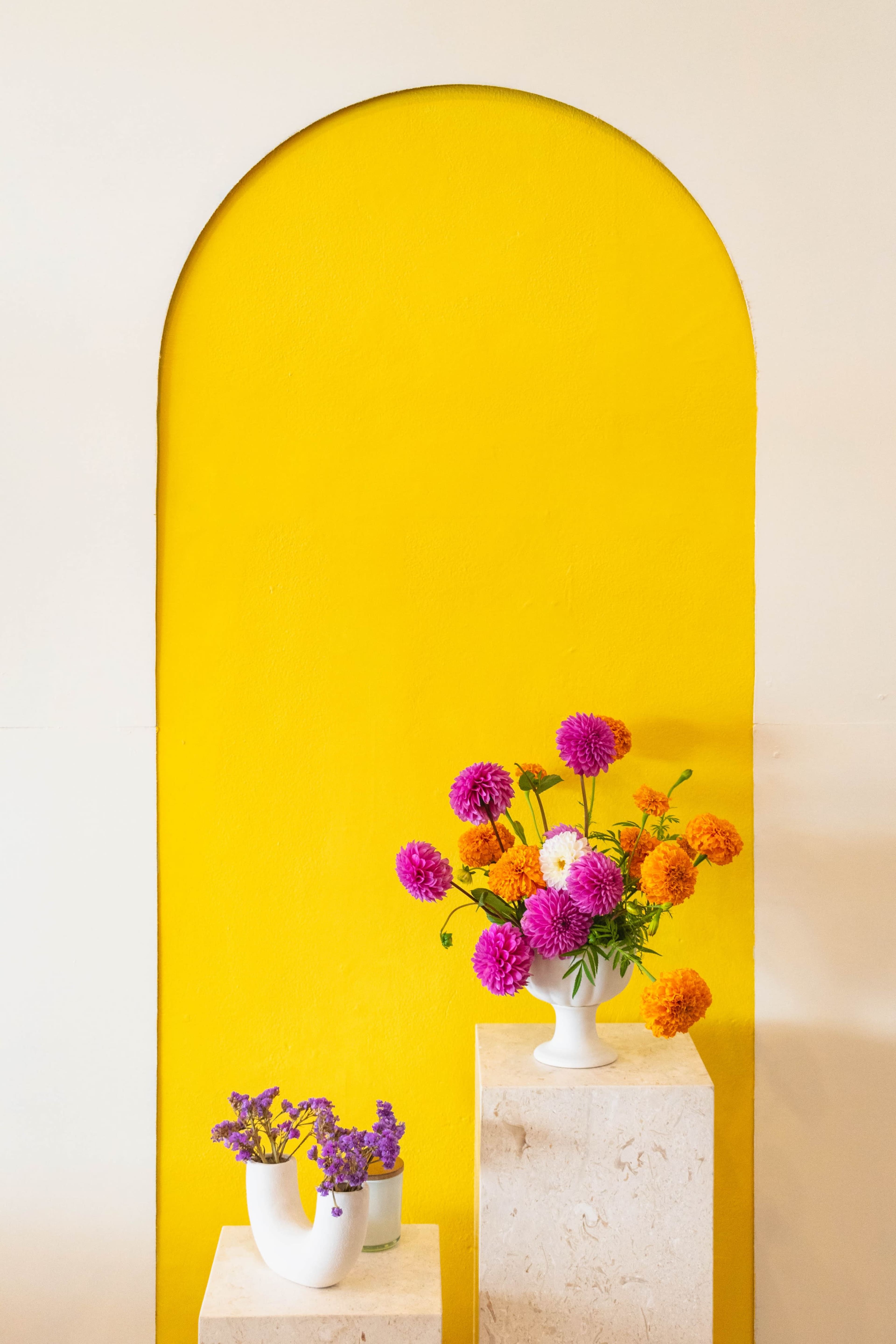 A vibrant yellow wall features an arch, with two stone pedestals displaying flower arrangements in white vases.
