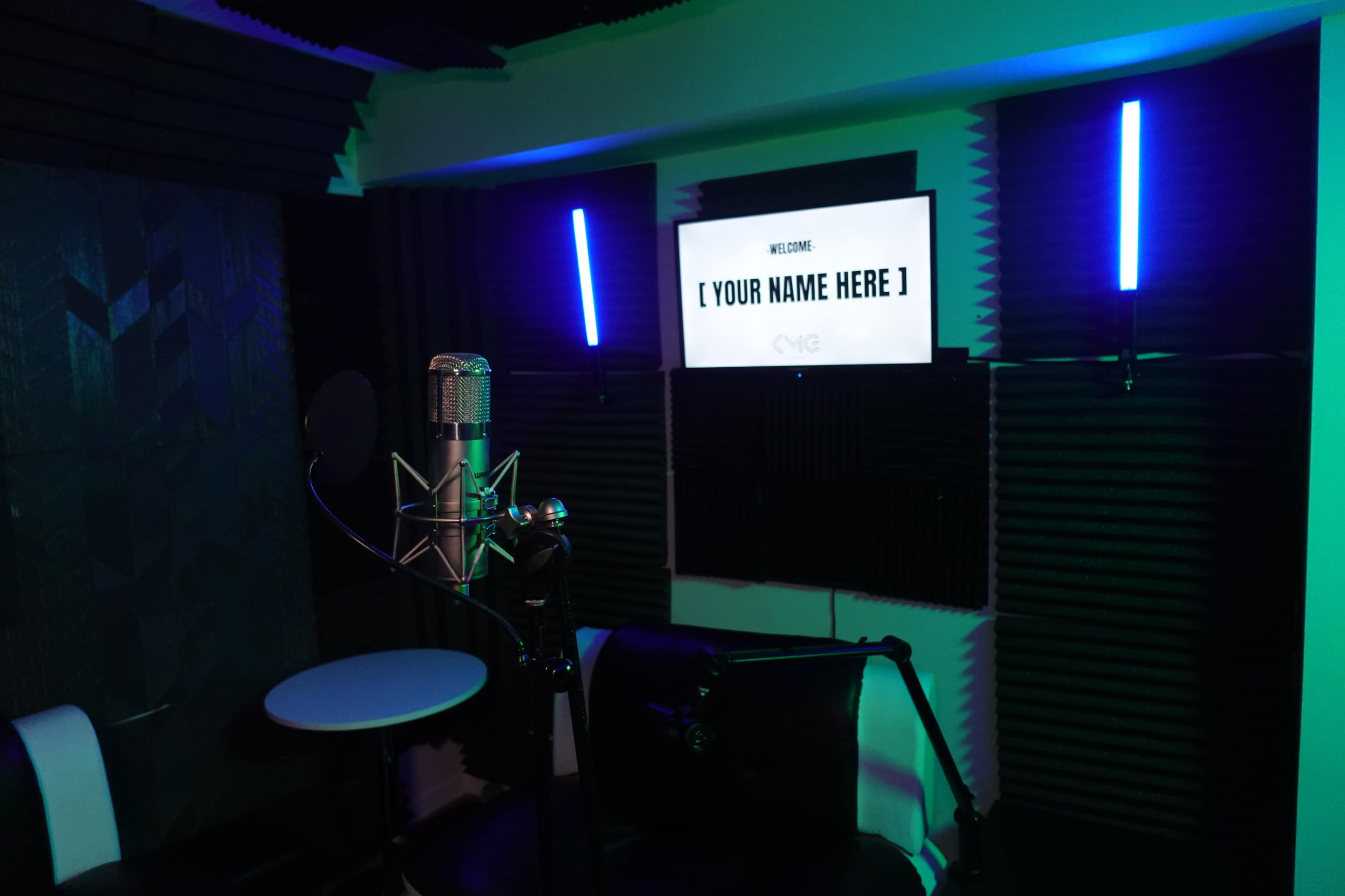 The image shows a recording studio with a microphone, a small table, and a screen displaying “[ YOUR NAME HERE ]” on the wall.