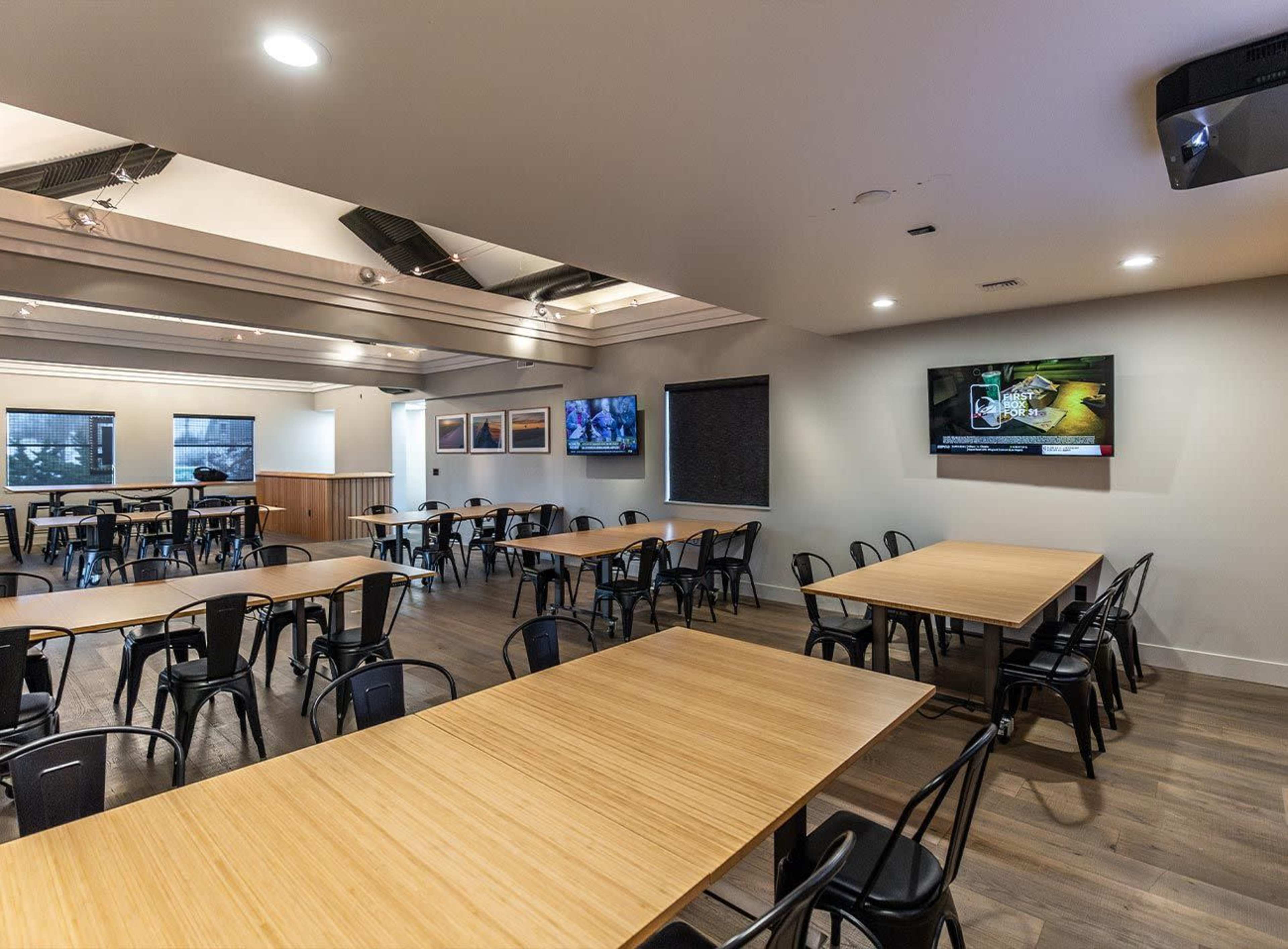 A spacious dining area with multiple wooden tables and black chairs, featuring a television mounted on the wall.