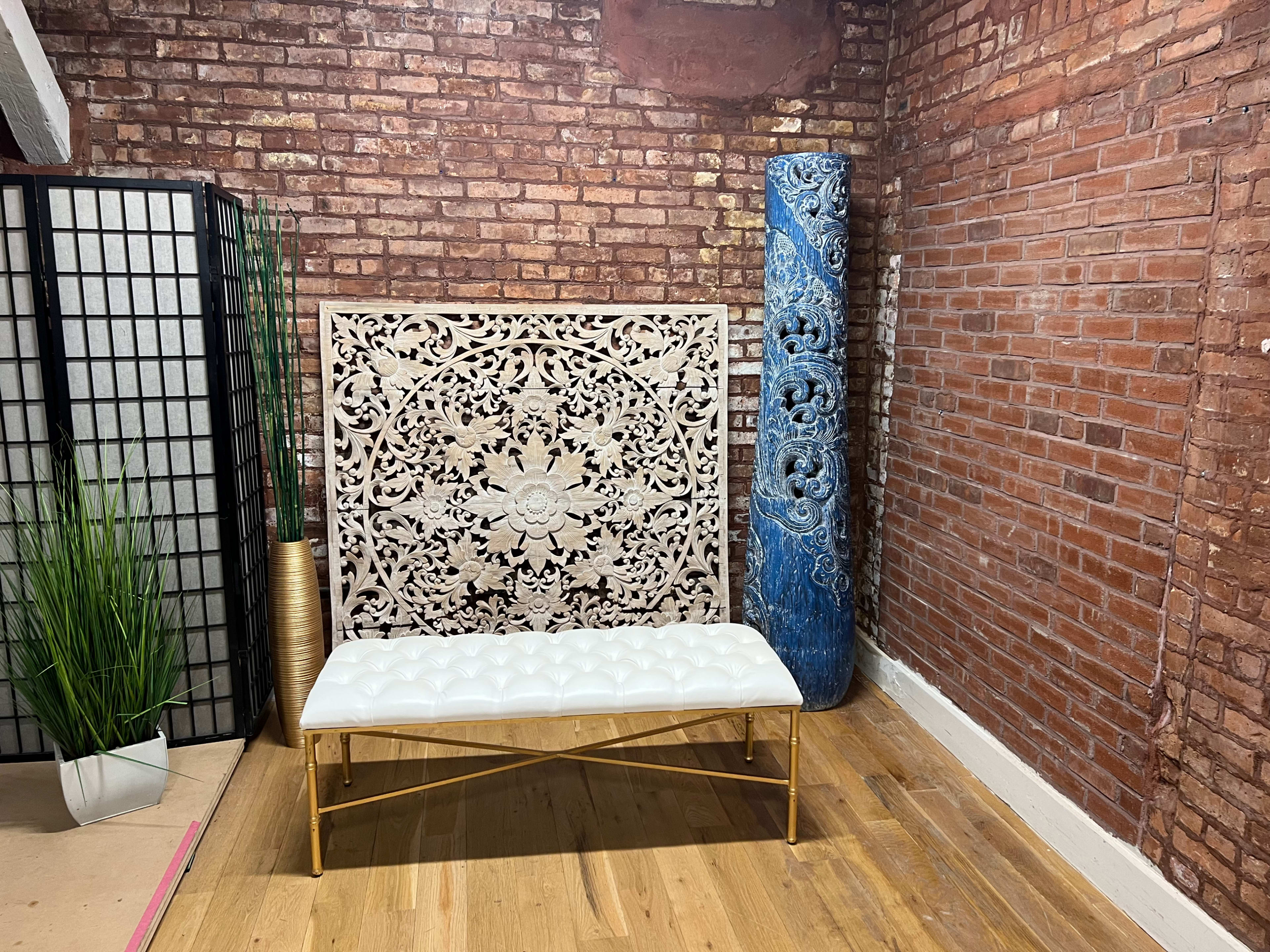 The image shows a decorative room with a white tufted bench, a carved backdrop, a tall blue decorative column, and a potted plant against a brick wall.
