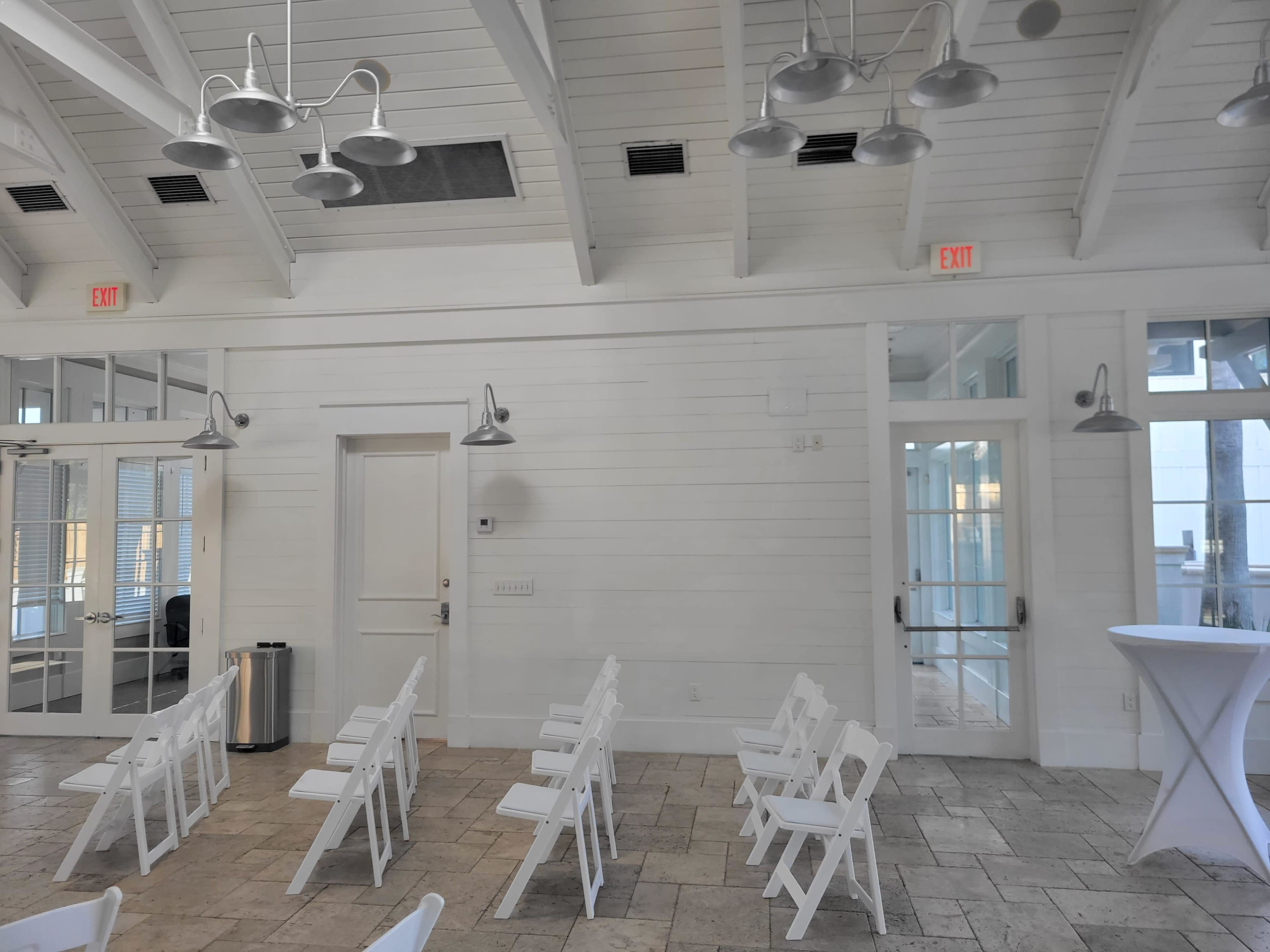 A bright interior space with white walls and a few rows of folding chairs arranged facing a doorway.