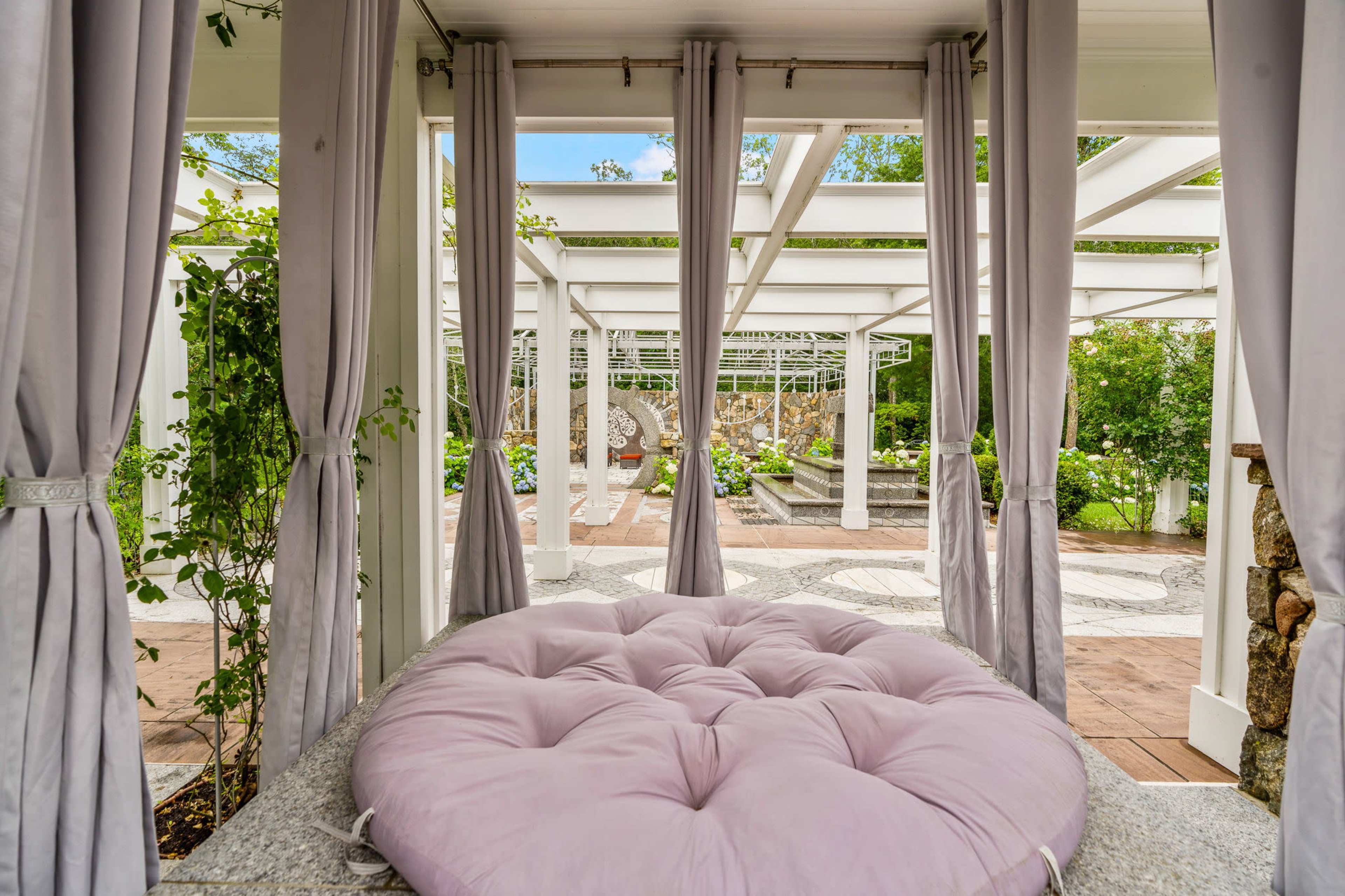 A spacious outdoor area is framed by curtains, showcasing a large circular cushion and a decorative stone path in the background.